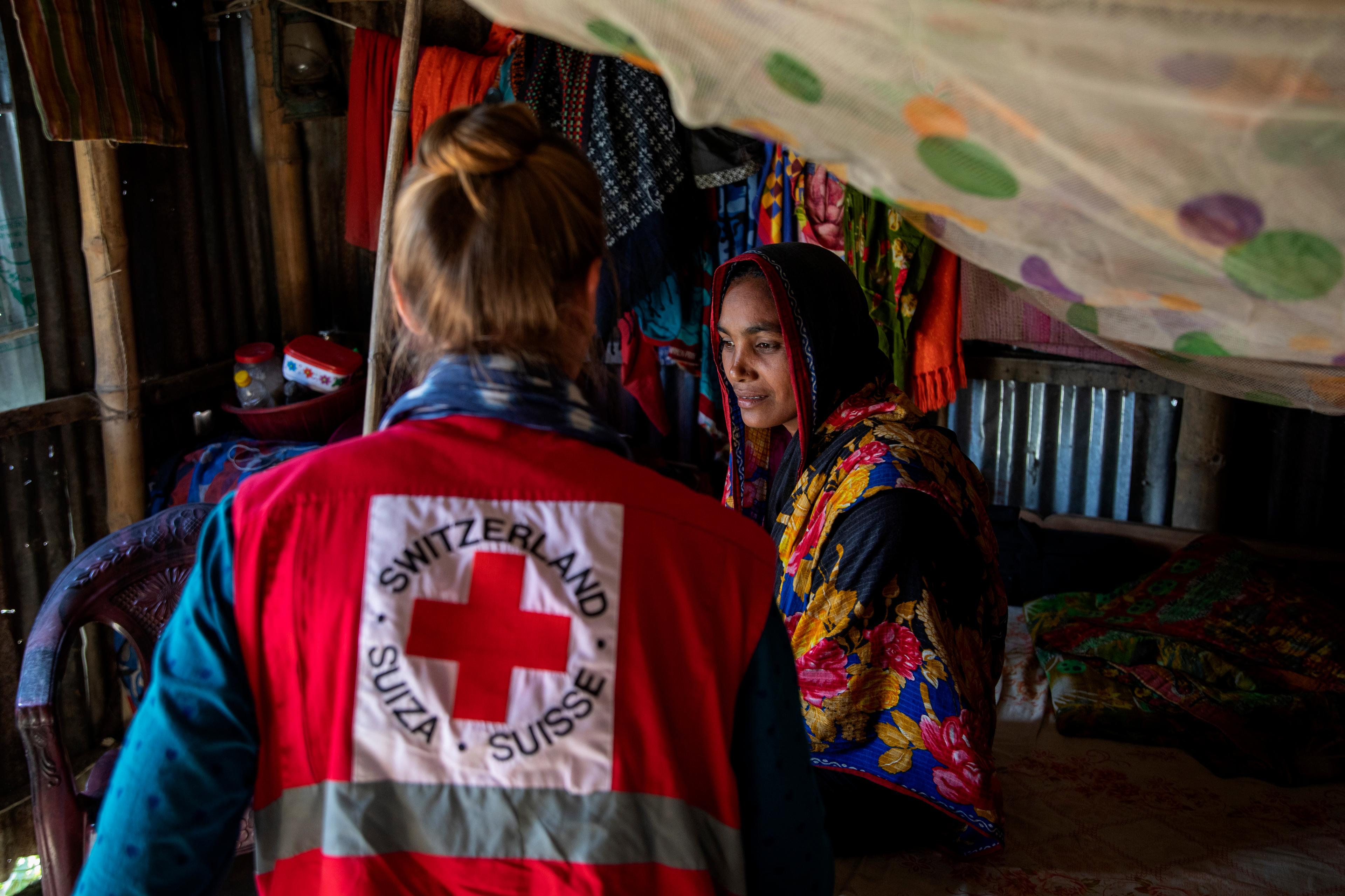 Zwei Frauen sitzen beeinander. Sie befinden sich in einer Wellblechhütte. Eine Frau trägt ein Tuch mit Blumen über Kopf und Oberkörper. Die zweite trägt eine rote Weste mit dem Schweizerischen Roten Kreuz auf dem Rücken.