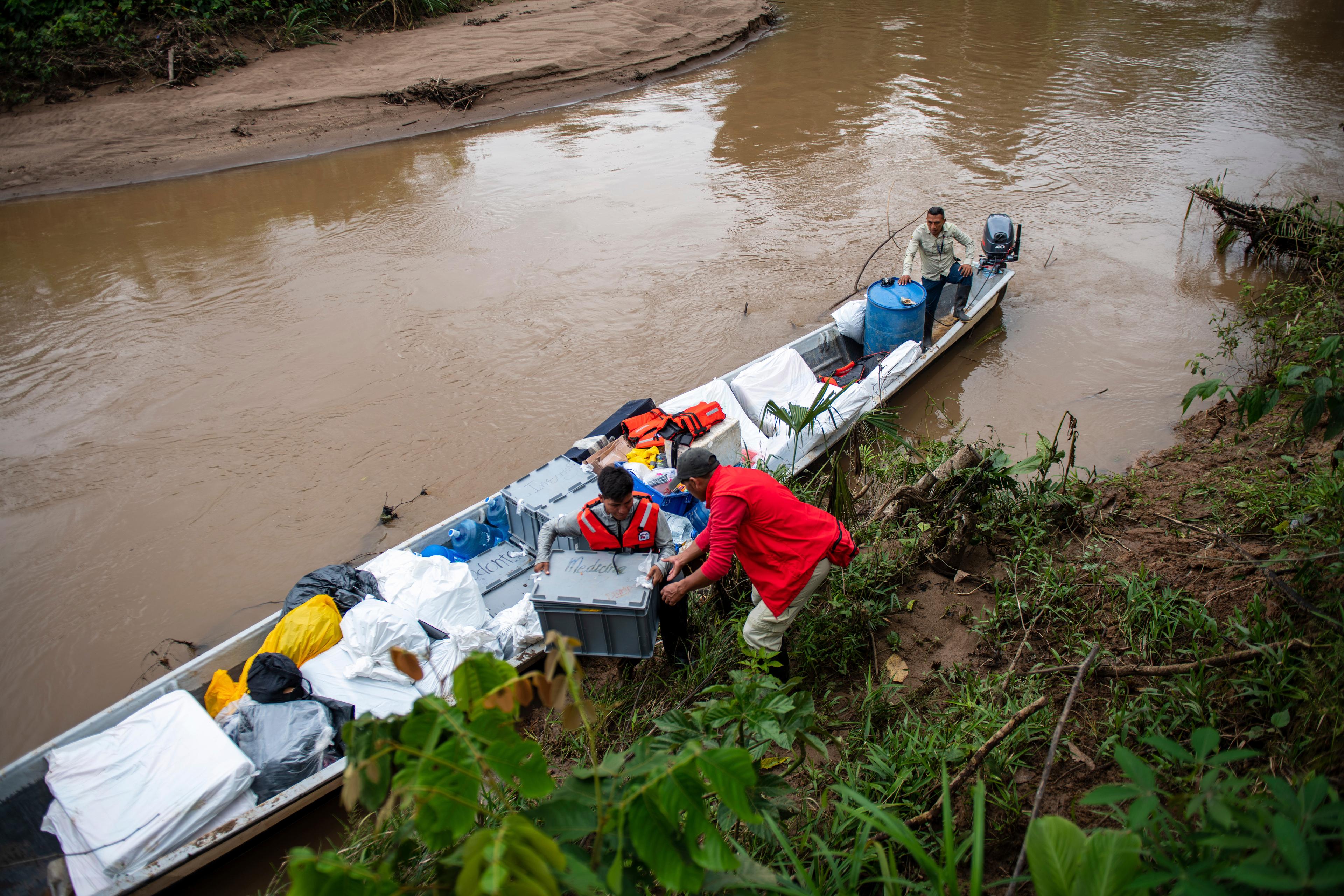 Alcune persone scaricano delle attrezzature da una semplice imbarcazione di legno attraccata in riva a un fiume.