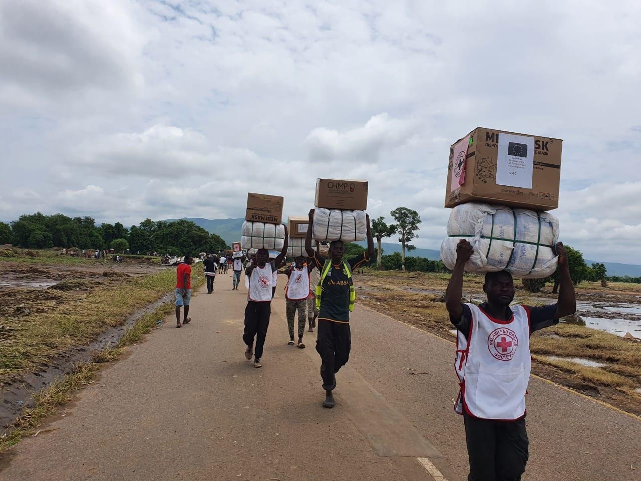 Plusieurs hommes des t-shirt de la Croix-Rouge portent des paquets au-dessus de leur tête. Ils avancent sur une route qui est entourée par des champs innondés.