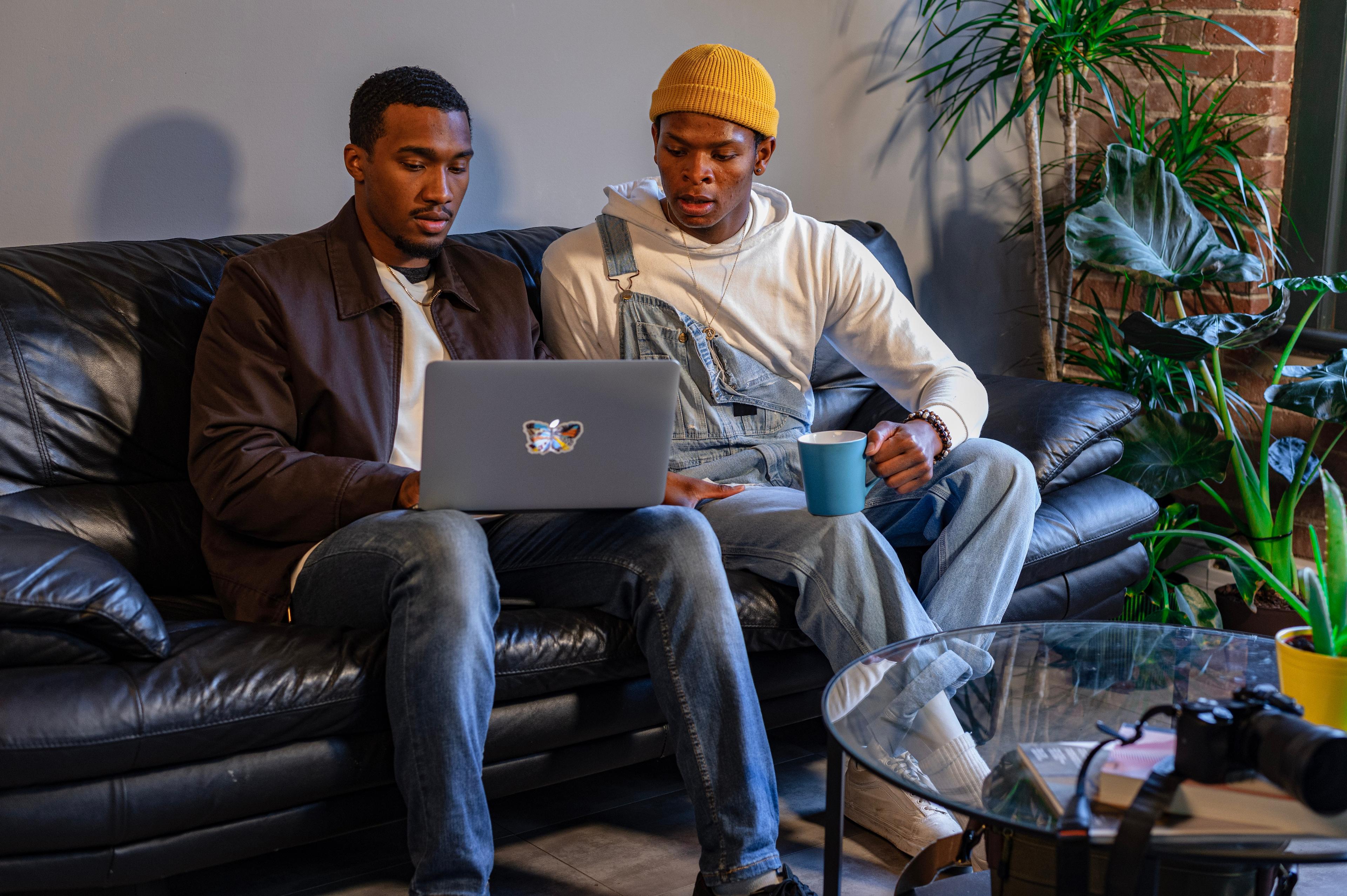 Due uomini sono seduti su un divano in un'atmosfera familiare, uno ha un computer portatile sulle ginocchia; entrambi guardano lo schermo.