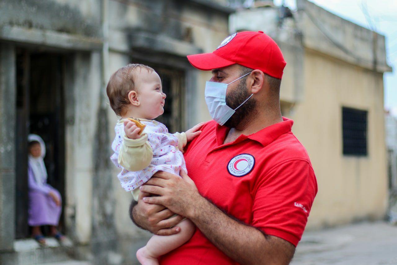 Un collaborateur du Croissant-Rouge arabe syrien tient un bébé dans ses bras, ils se sourient.