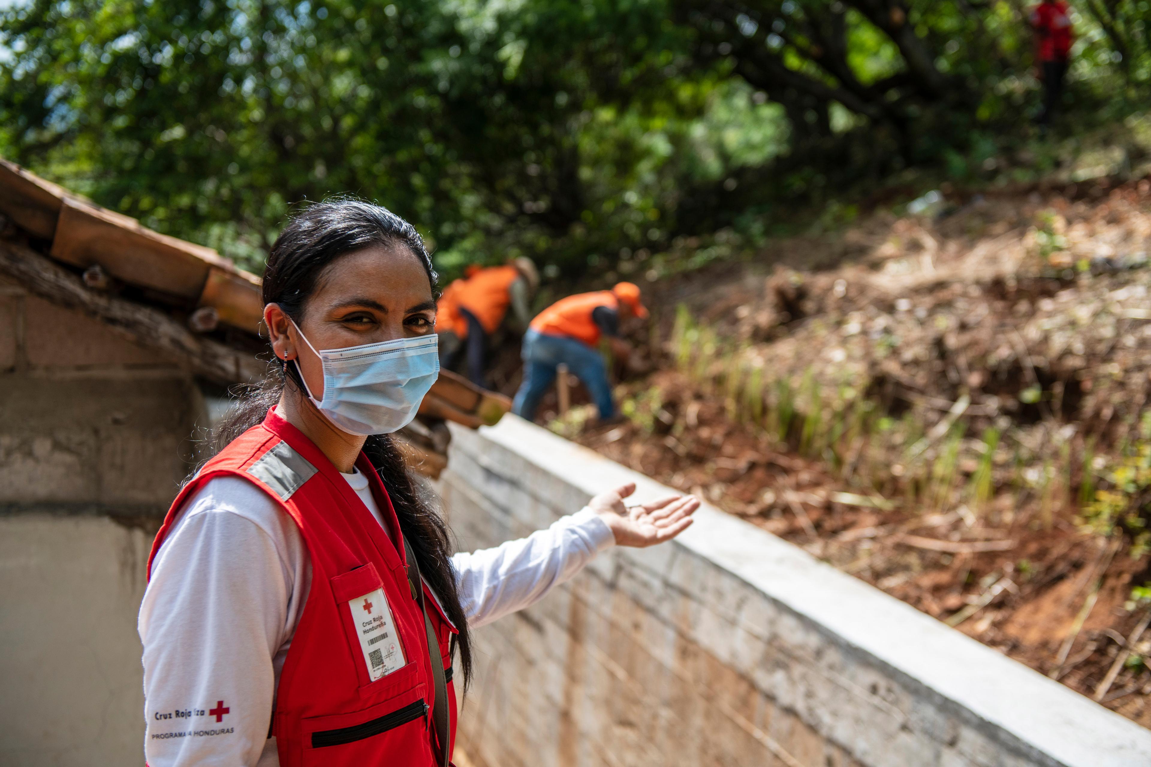 A woman wearing a red waistcoat and sanitary mask looking at the camera, in the back two farm workers.