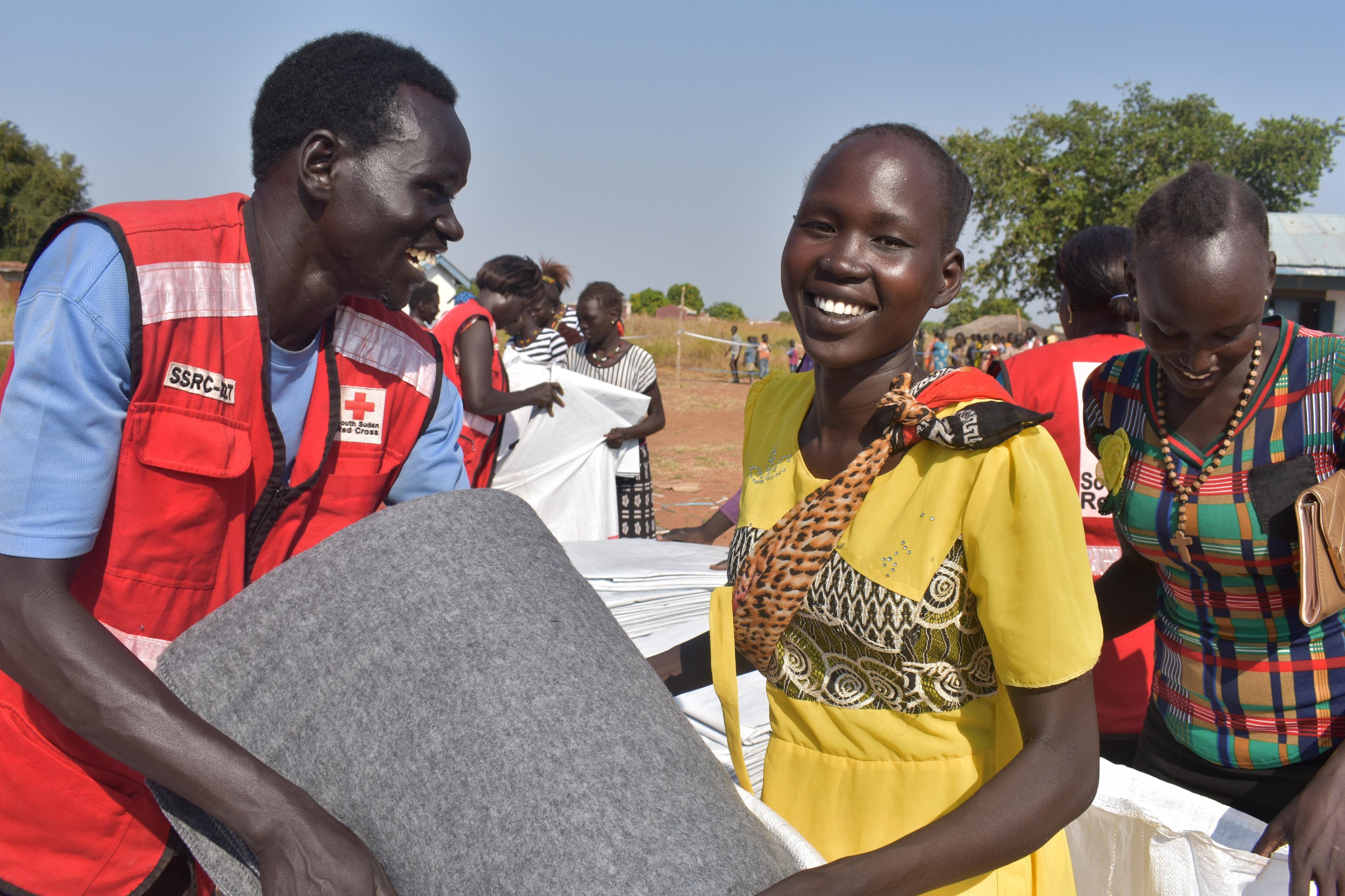 A man in a Red Cross vest smilingly hands over a blanket to a woman who also has a smile on her face. More such handovers take place in the background.  