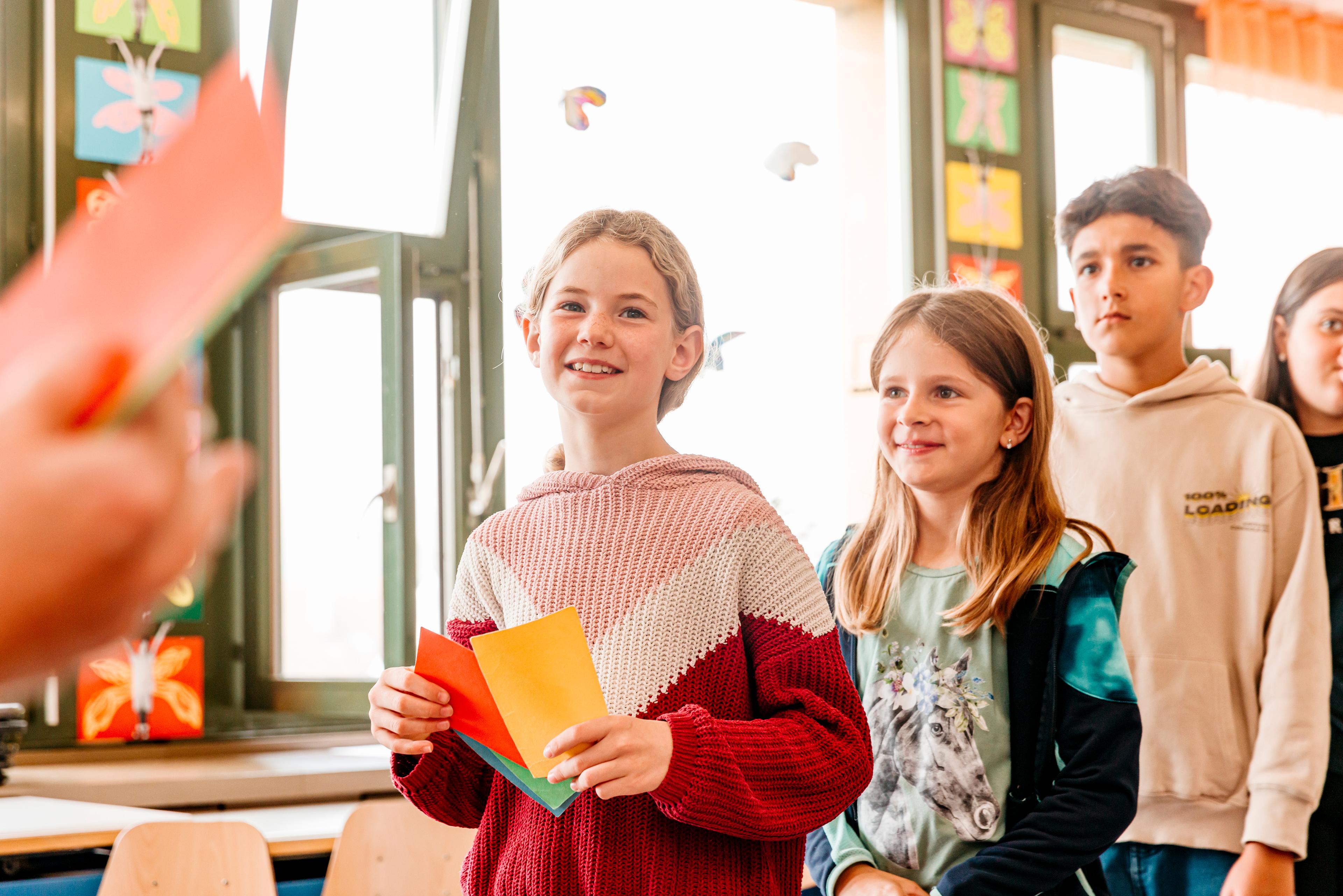 Plusieurs enfants sont en colonne dans une salle de classe et participent à un exercice de l’atelier chili. La fille qui se trouve en tête tient dans la main des cartes de couleur verte, jaune, rouge et bleue, qu’elle peut lever en l’air. 