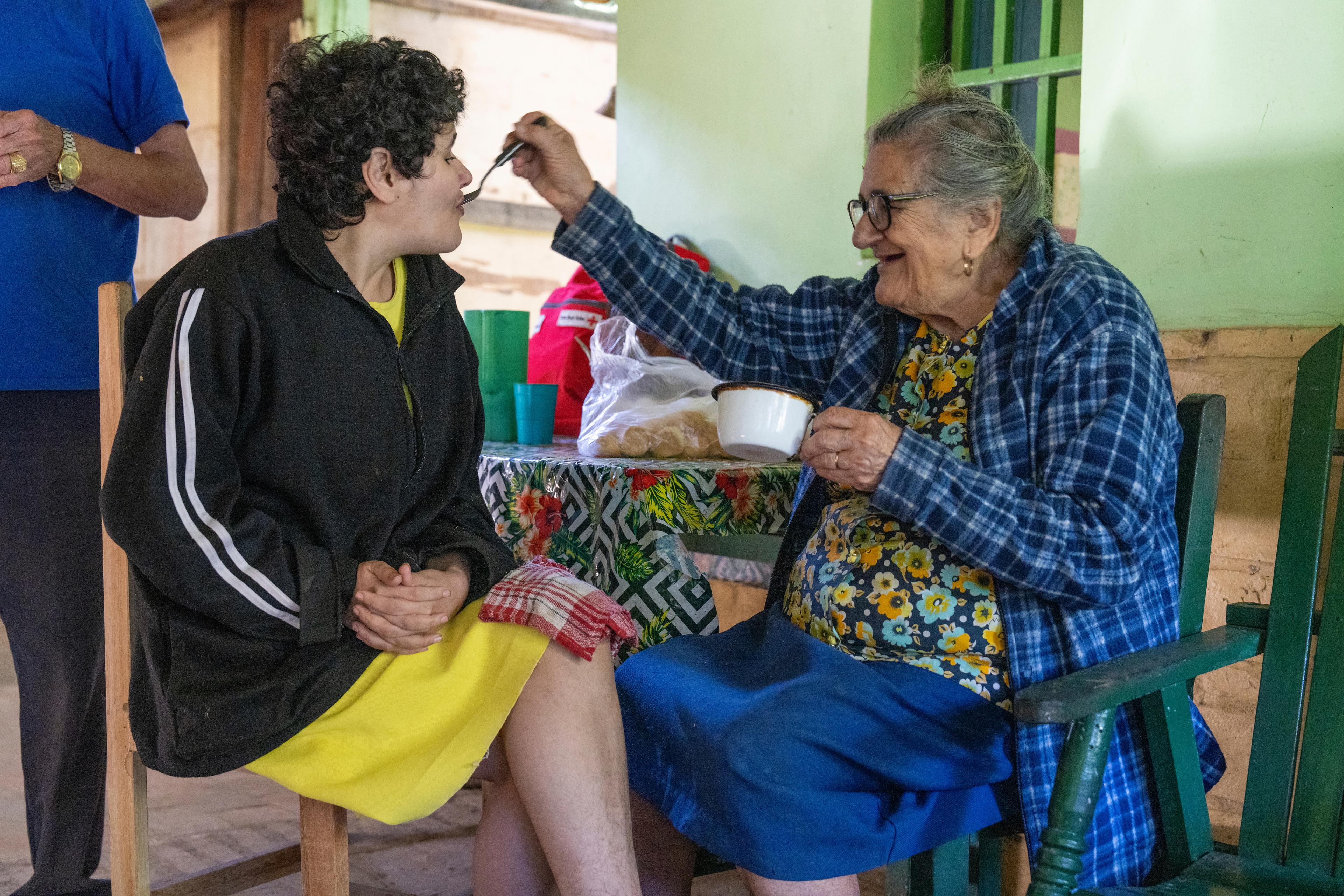 Une femme âgée donne à manger à sa fille; toutes deux sont attablées dans une cuisine.