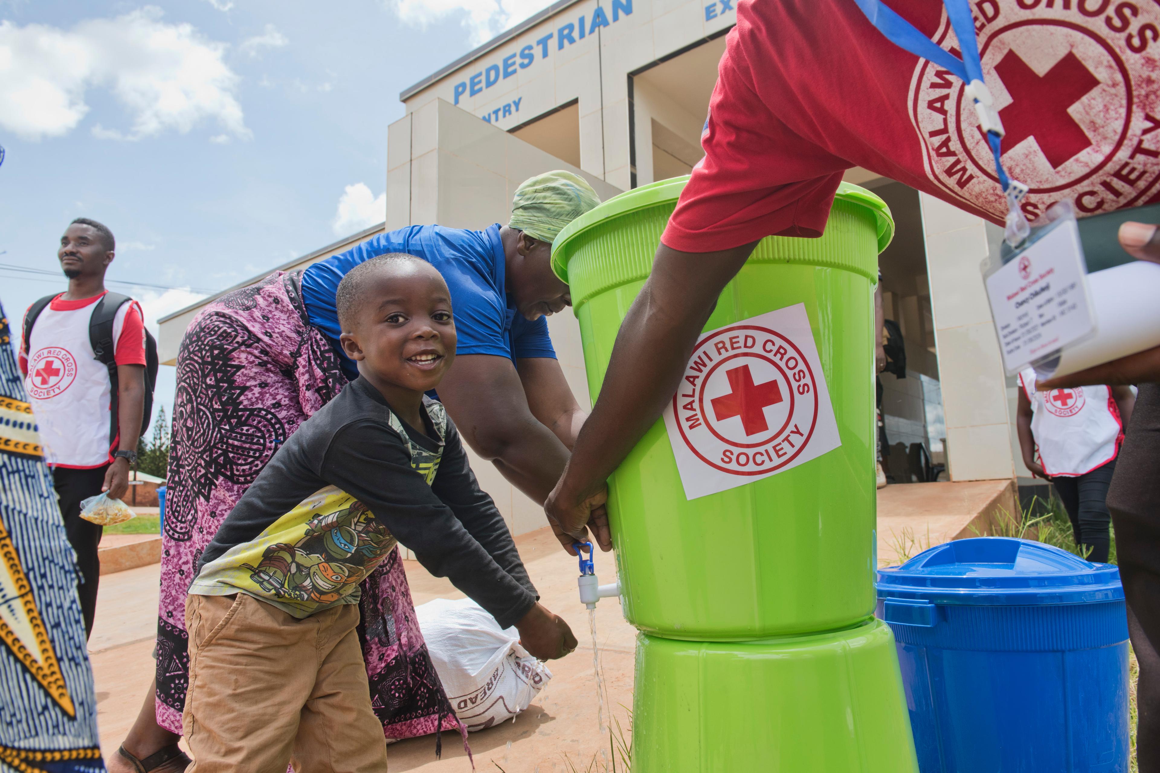 Ein kleines Kind wäscht sich die Hände aus einer Plastikschüssel, auf der ein großer Aufkleber des Roten Kreuzes von Malawi zu sehen ist. Eine erwachsene Person, deren Kopf nicht zu sehen ist, betätigt den Wasserhahn. Auf dem T-Shirt der erwachsenen Person ist das gleiche Logo des Malawischen Roten Kreuzes zu sehen.