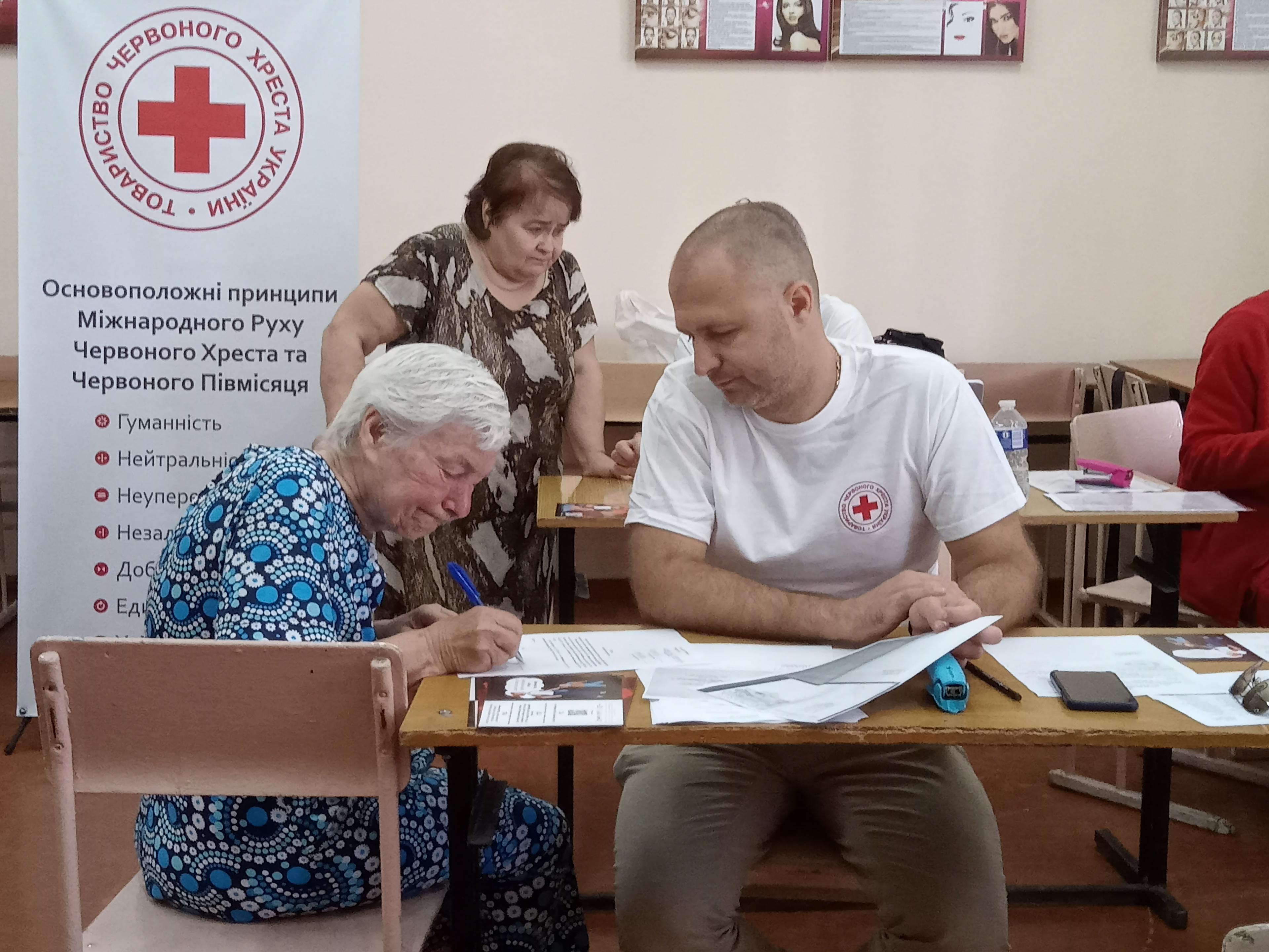 4.	Une femme âgée remplit des documents à une table. Un homme à ses côtés tient une partie des documents. Sur son t-shirt figure un logo de la Croix-Rouge. L’homme semble assister la femme pour remplir les documents.