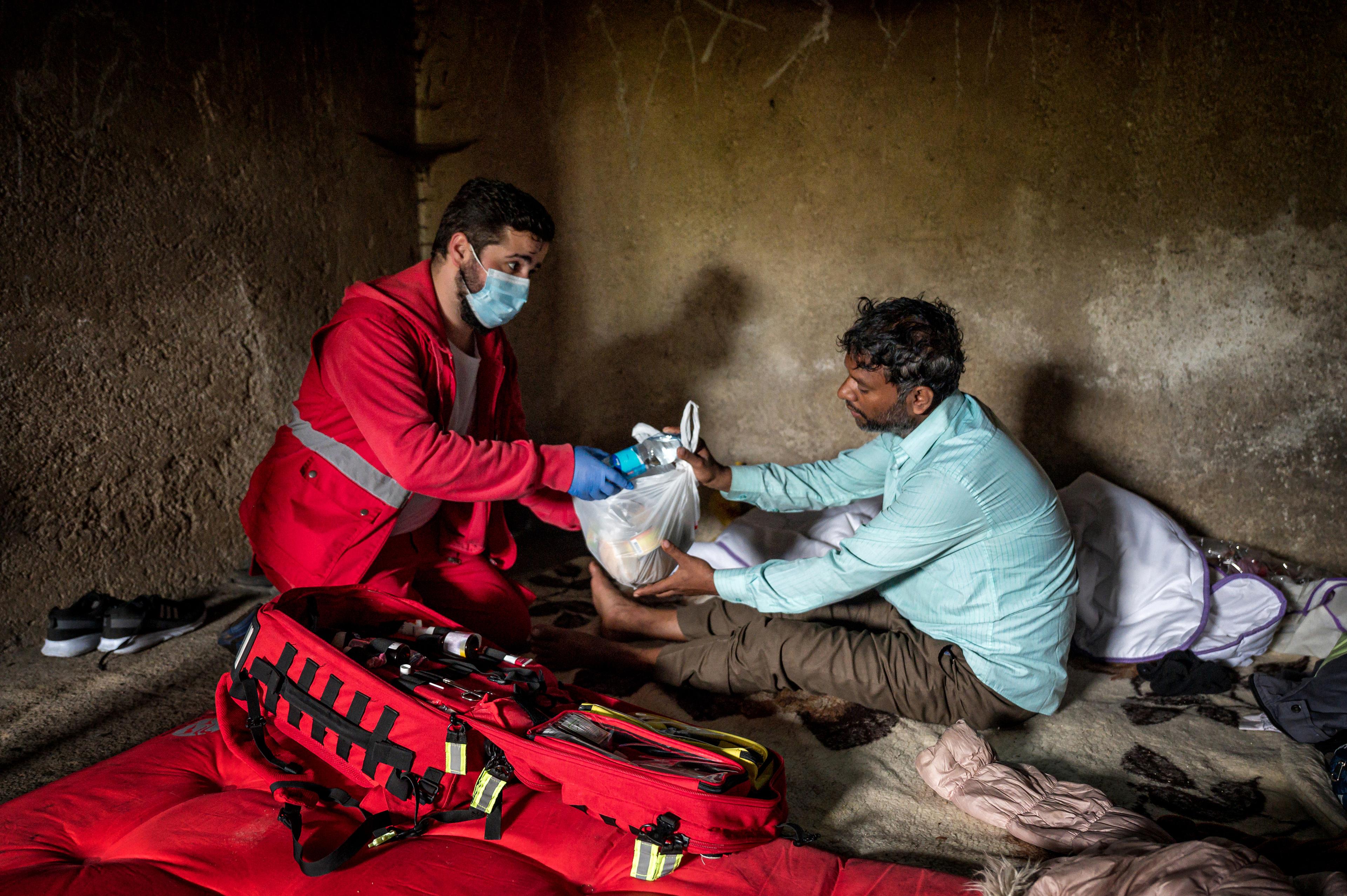 The image shows an employee of the Red Cross movement in a red uniform, handing over supplies to a seated man in a sparsely furnished room, depicting a field operation or humanitarian assistance context.