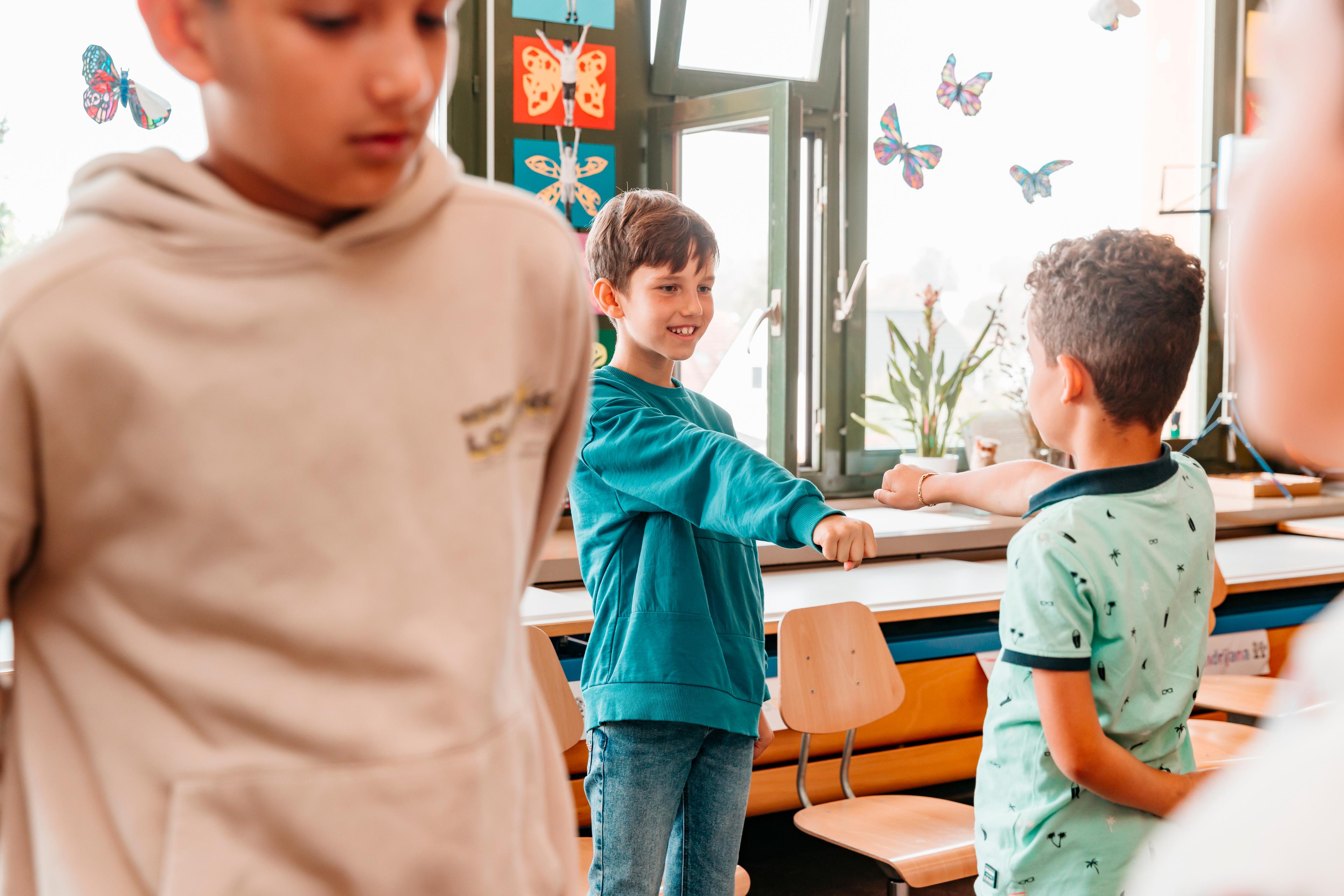 Zwei Kinder stehen sich in einem Klassenzimmer lächelnd gegenüber und strecken sich die Faust entgegen. Sie machen bei einer Übung vom chili Konflikt-Training mit. 