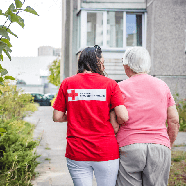 This picture shows a woman with a red t-shirt holding a woman with white hair by the arm. On a path next to a building. 