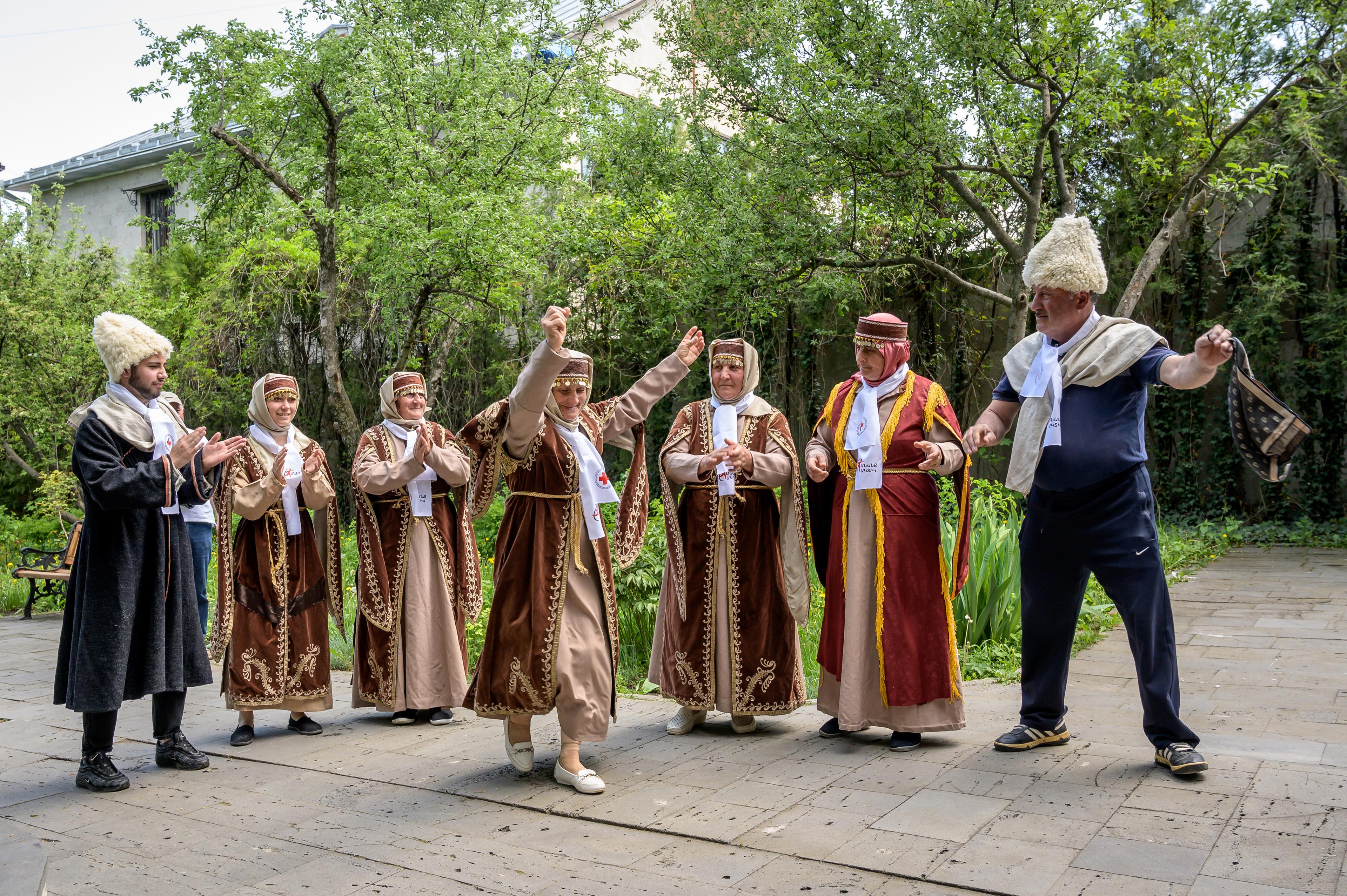 A picture showing a group of men and women dancing in traditional clothes. An area outside with trees.