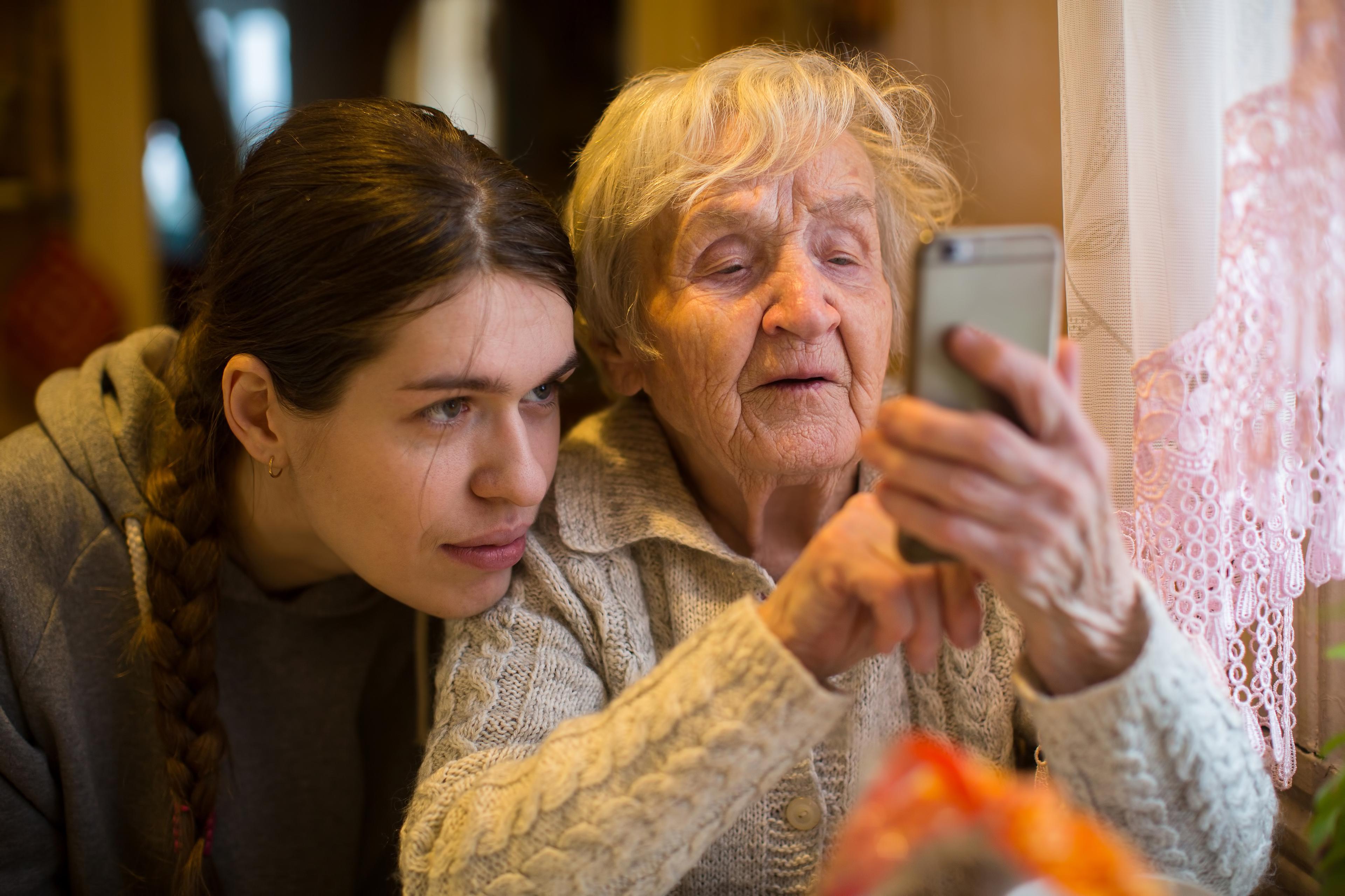 Das Gesicht eines jungen Mädchens ähnelt stark dem einer älteren Dame. Sie schauen gemeinsam auf den Bildschirm eines Mobiltelefons.