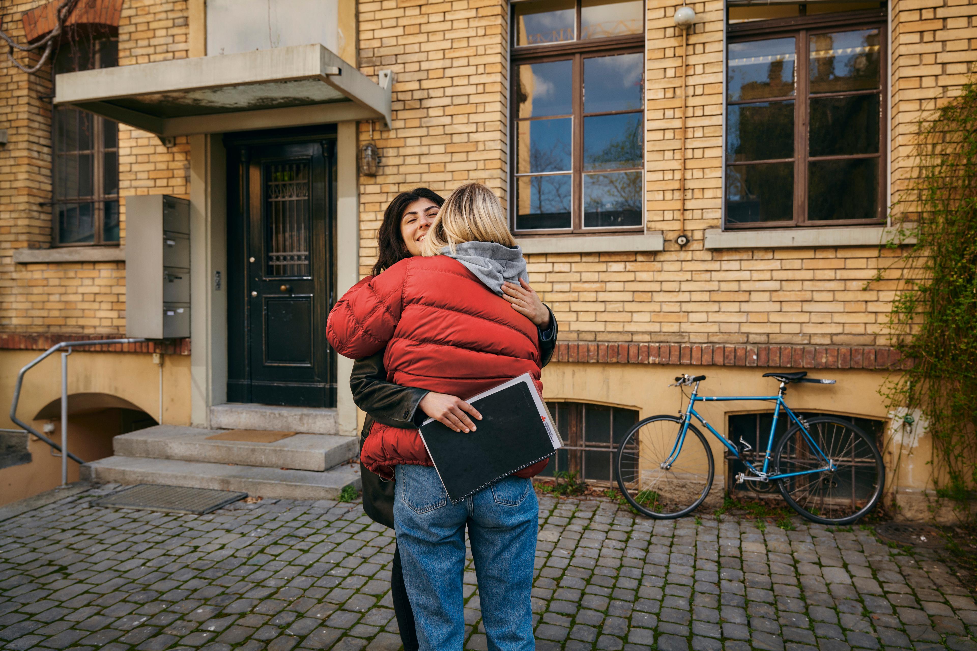 Eine Frau in roter Jacke mit dem Rücken zur betrachtenden Person umarmt eine lachende Frau, welche Dokumente in den Händen hält und die Umarmung erwidert. Die beiden stehen vor einem Hauseingang.