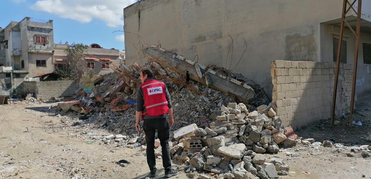 Un homme se tient devant les décombres de maisons. Il nous tourne le dos et porte un gilet rouge orné du logo de la Fédération internationale des Sociétés de la Croix-Rouge et du Croissant-Rouge. Le ciel est bleu, et il fait beau. 