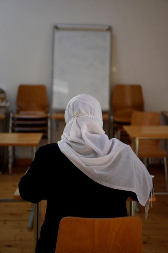 Una donna fotografata di spalle, seduta al banco di un'aula, che guarda una lavagna a fogli mobili davanti a sé.