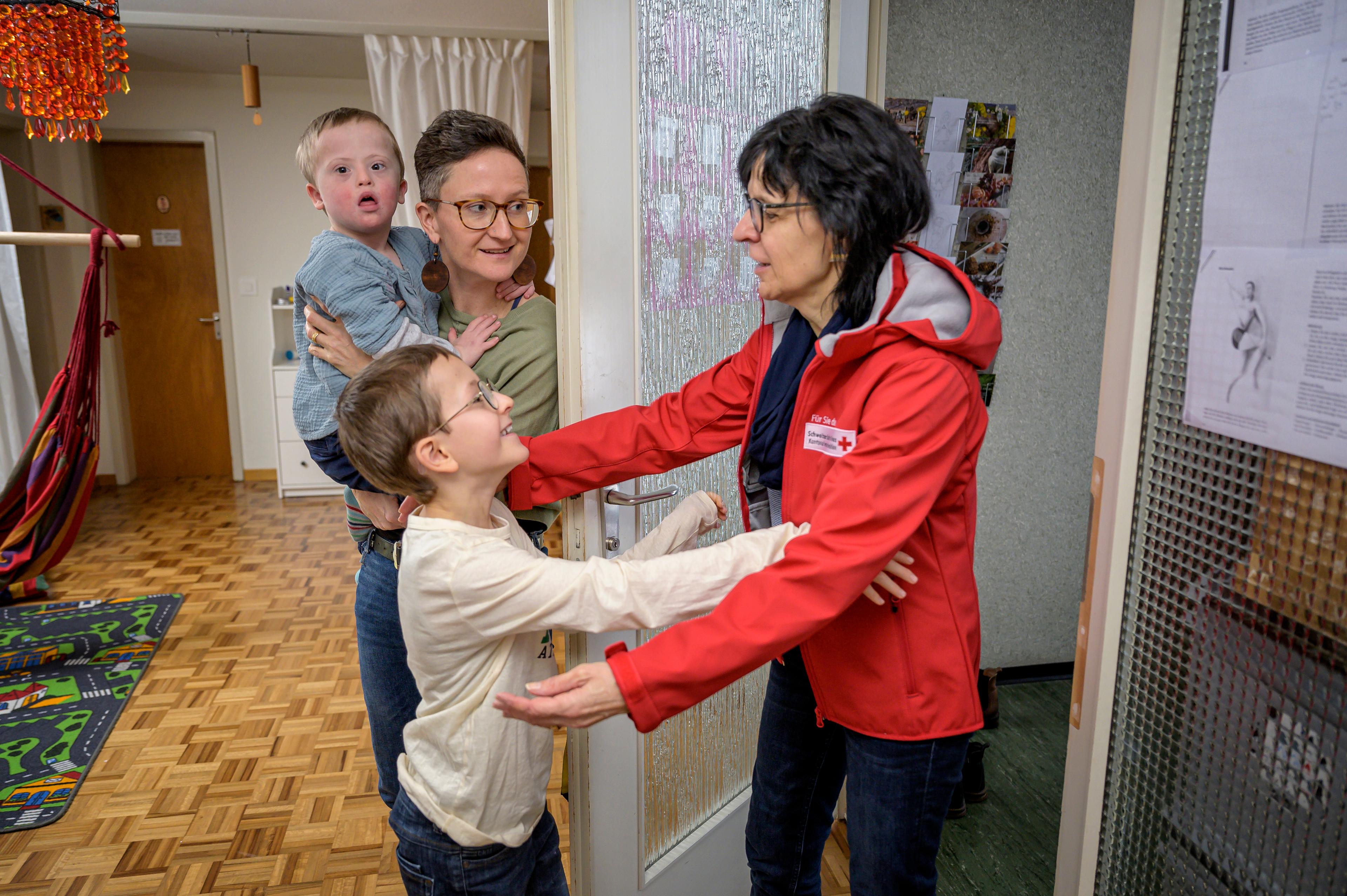 Une mère avec un petit enfant atteint de trisomie 21 dans les bras. Un enfant d'environ 10 ans salue la femme portant une veste rouge et le logo de la Croix-Rouge.