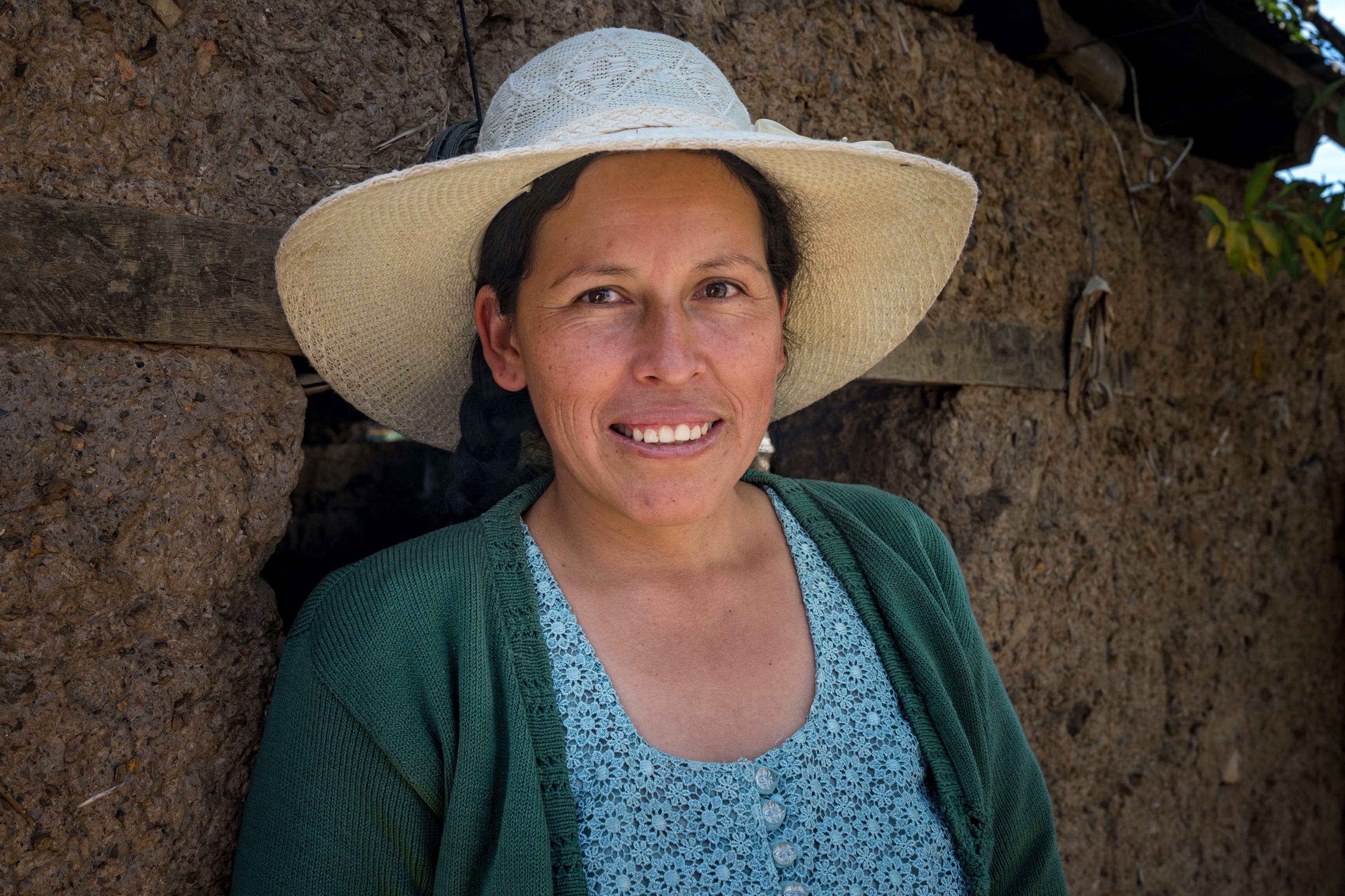 A smiling woman with a white hat stands before a stone wall.