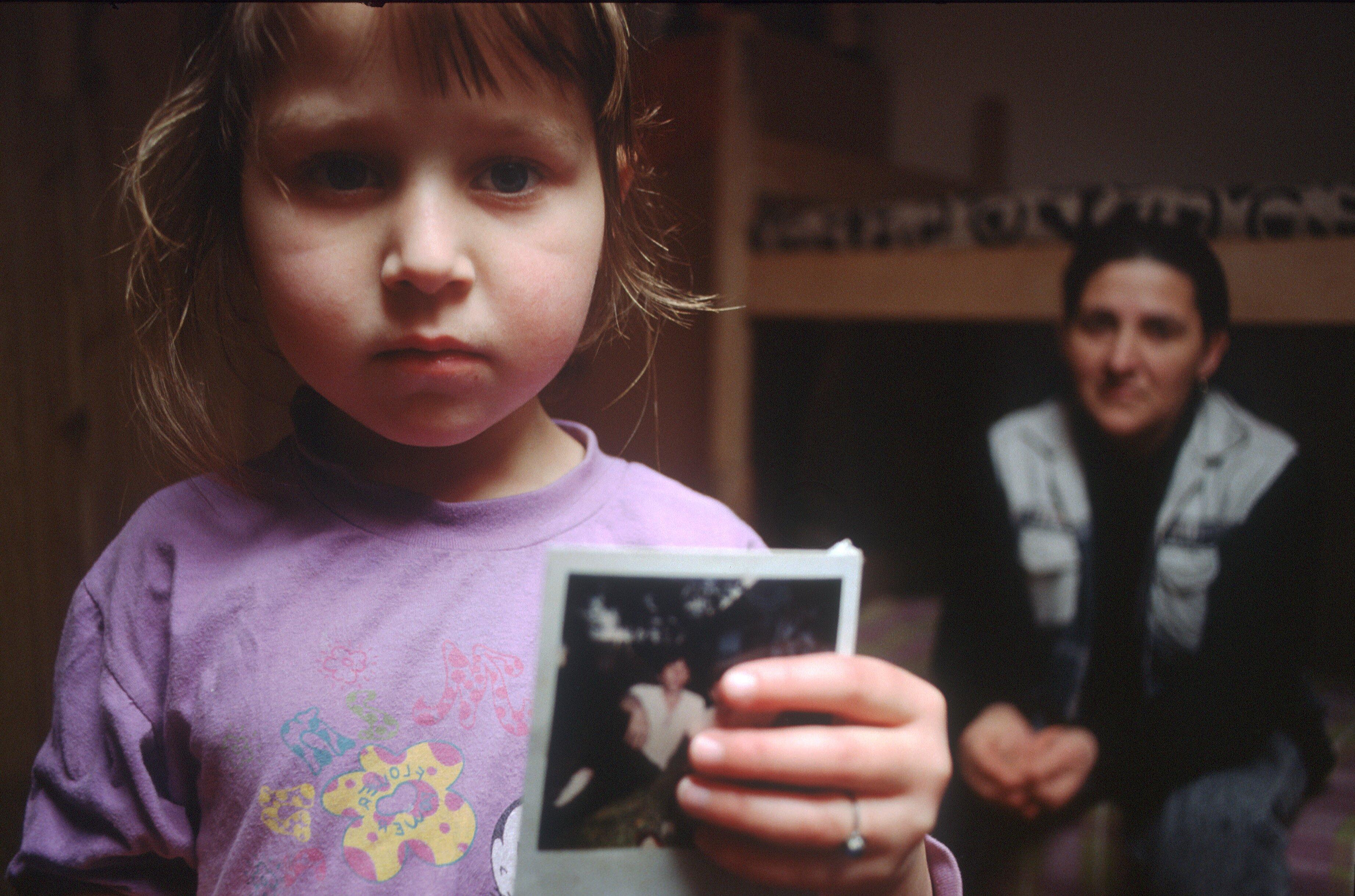 A young girl with a melancholic gaze gently holds a photograph in a bedroom. Behind her, an adult woman sits thoughtfully on the edge of a bed, enveloped in an atmosphere of nostalgia.