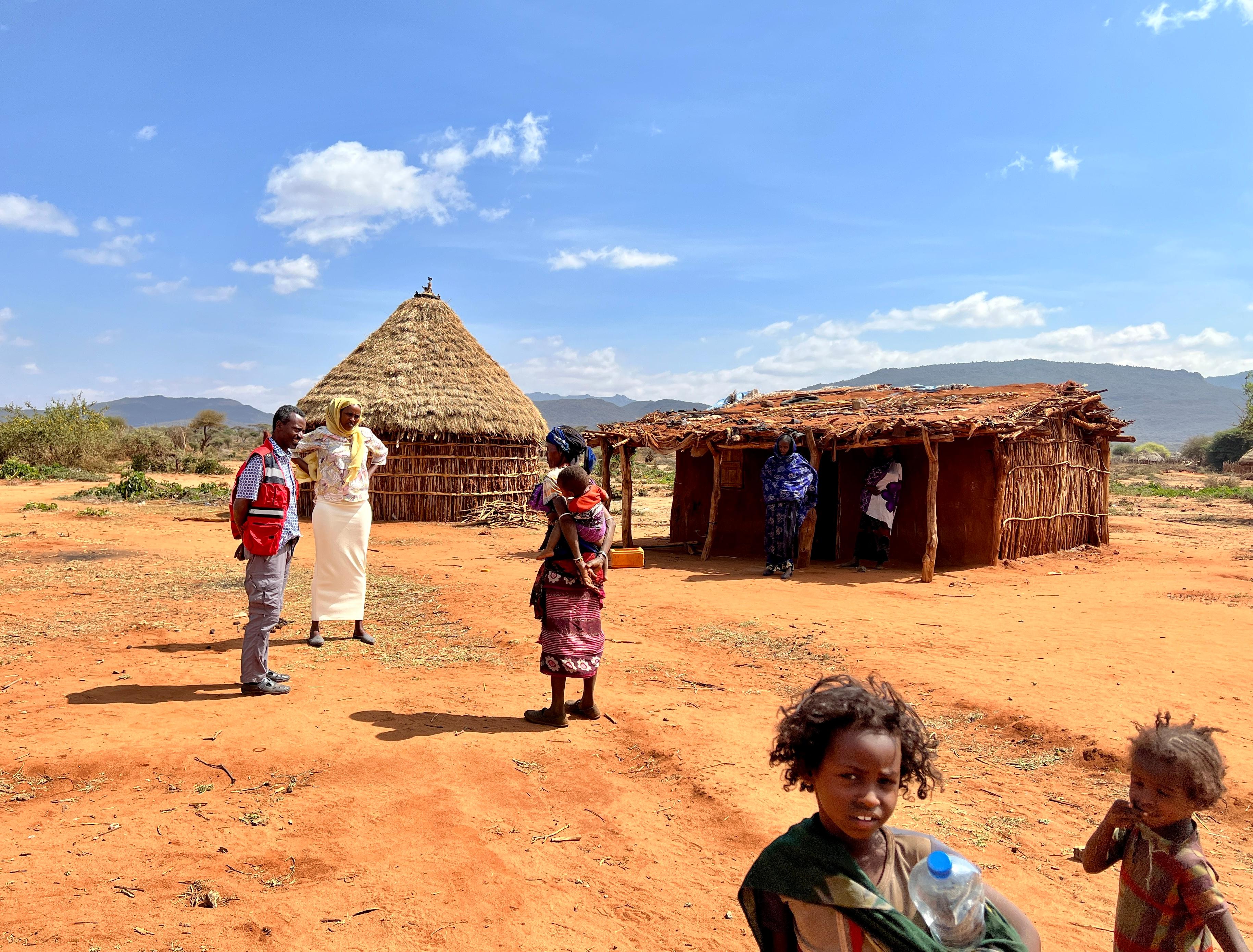 Eine Landschaft, die zwei Häuser aus Stroh und anderen Materialien zeigt. Davor befinden sich mehrere Personen, unter anderem auch ein Helfer des Roten Kreuzes und Kinder. 