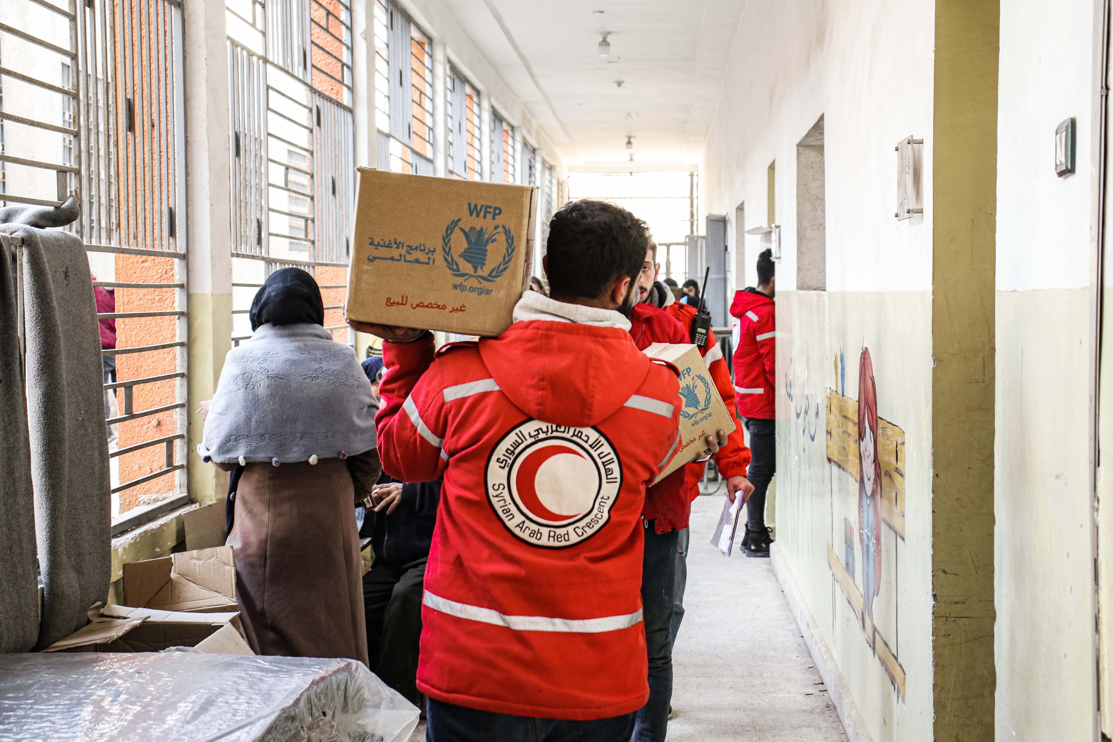 Un homme vêtu d’une veste aux couleurs du Croissant-Rouge marche dans un couloir en portant sur son épaule une caisse contenant des biens de secours.