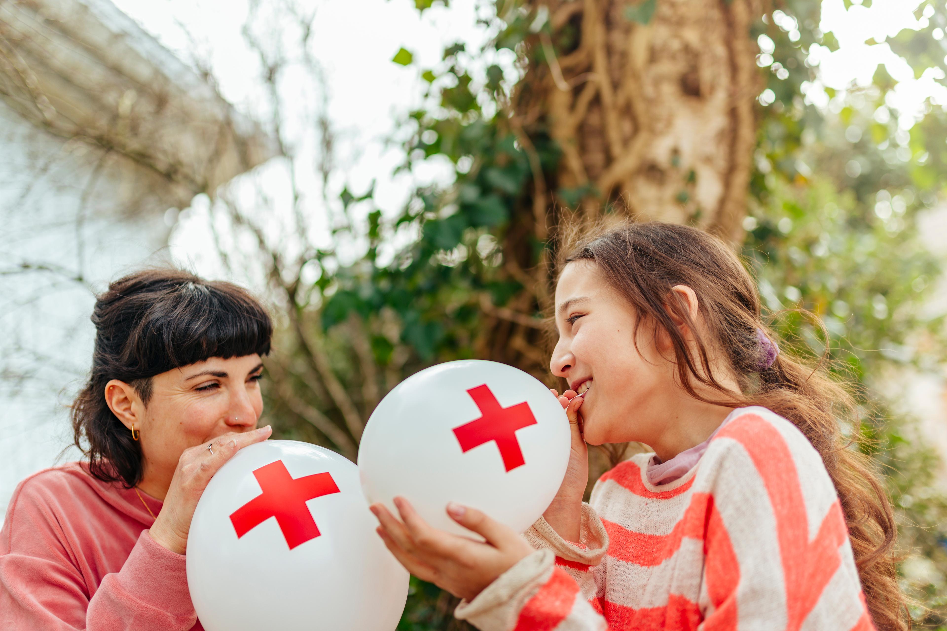 Dans un jardin, une mère et sa fille gonflent des ballons ornés d’une croix rouge.
