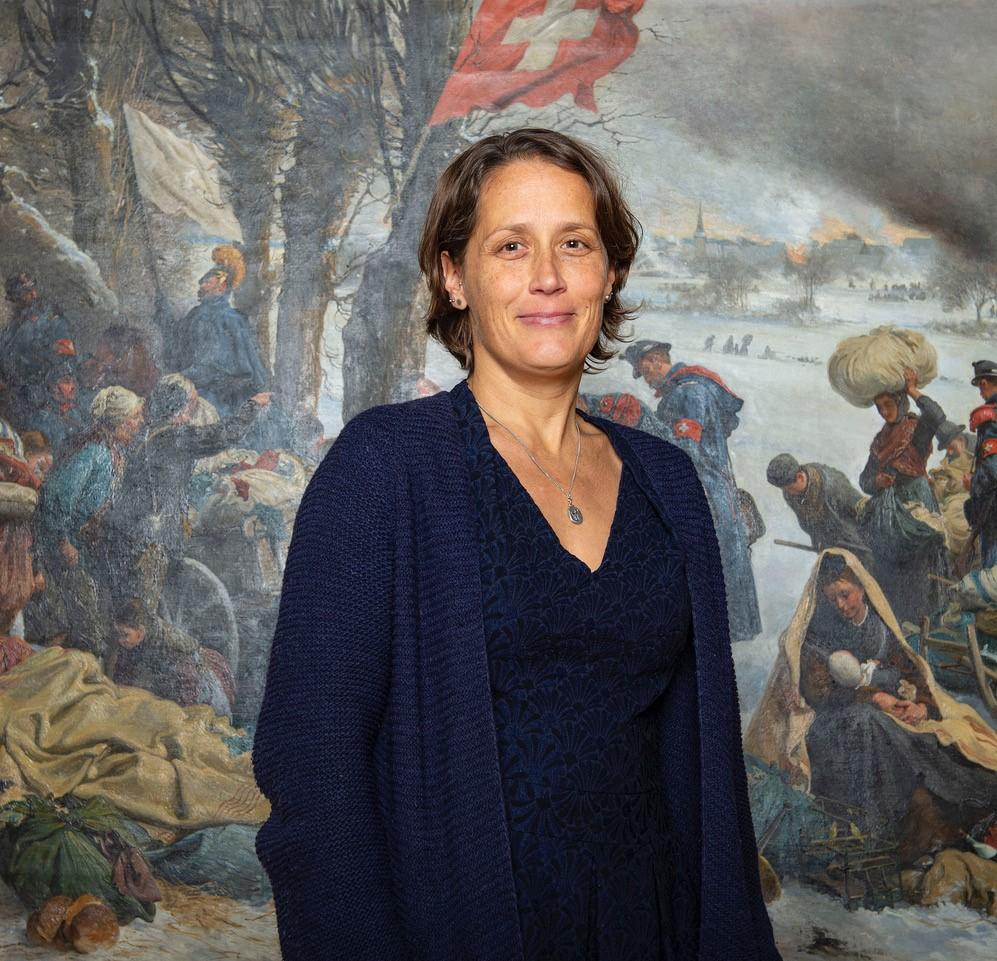 Nicole Windlin, Leiterin des Suchdienstes des Schweizerischen Roten Kreuzes,  steht vor einem beeindruckenden historischen Wandgemälde des Schweizerischen Roten Kreuzes darstellt.