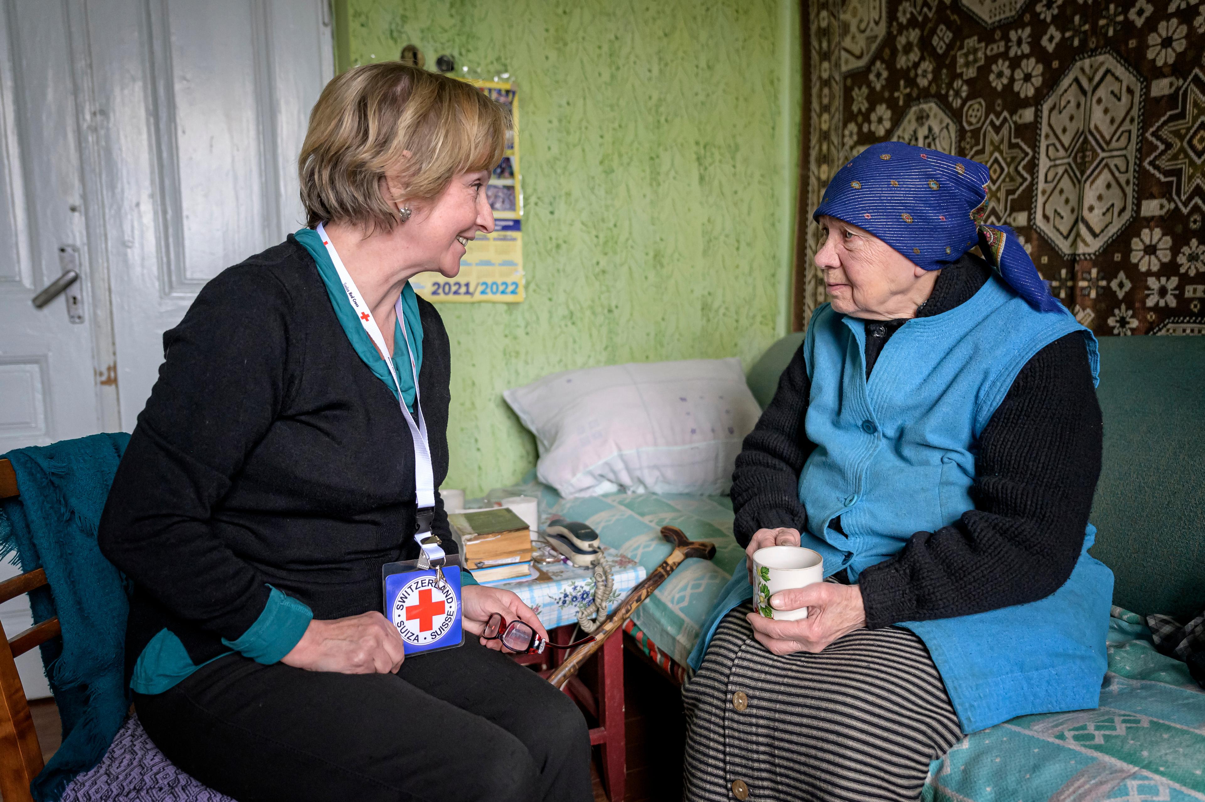 Zwei Frauen in einem Zuhause. Eine ältere Frau sitzt auf dem Bett, ihr gegenüber auf einem Stuhl Lidja Negid. Sie lächeln sich an.