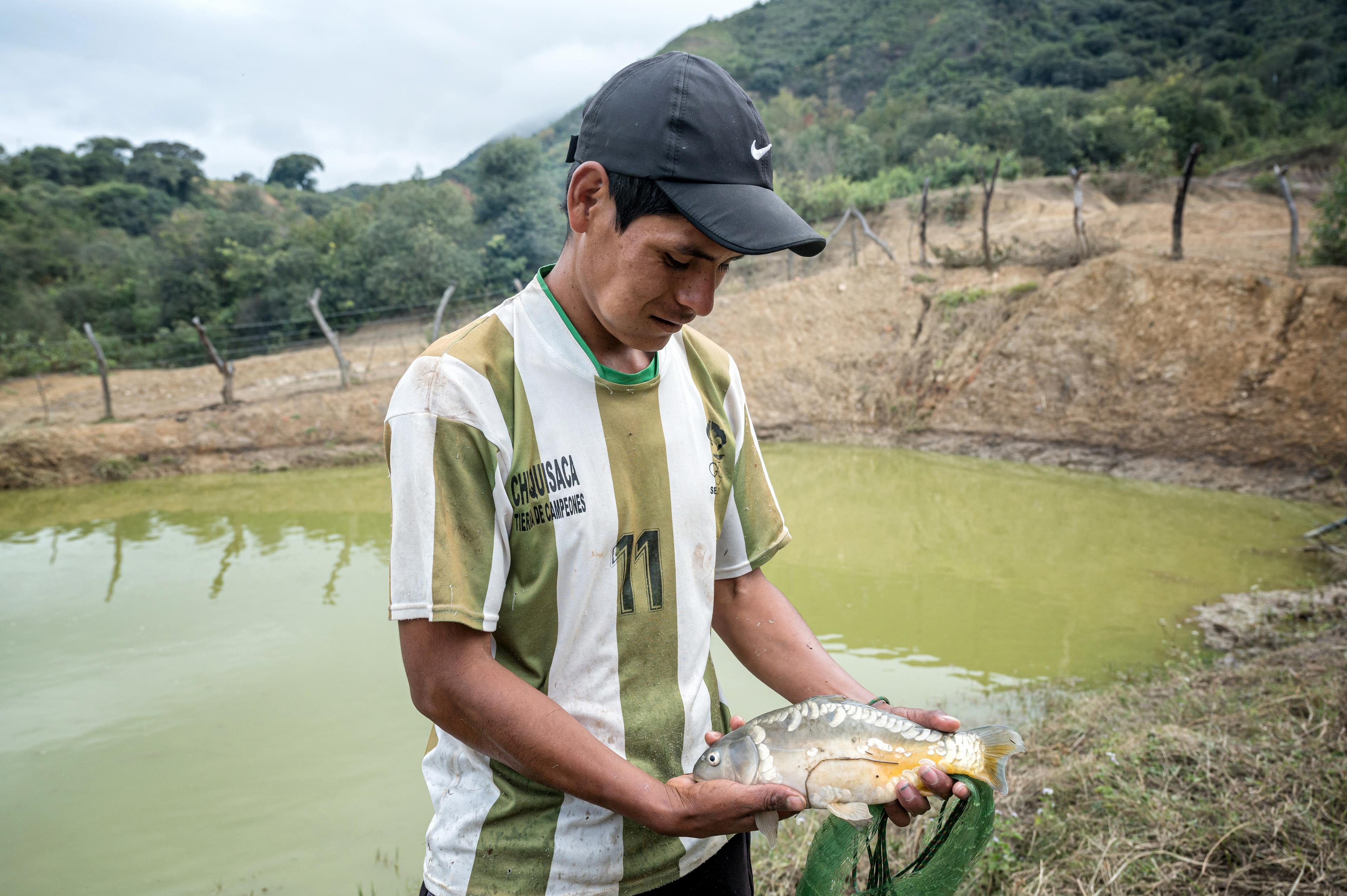 Un homme debout devant un étang tient un poisson dans ses mains et le regarde.