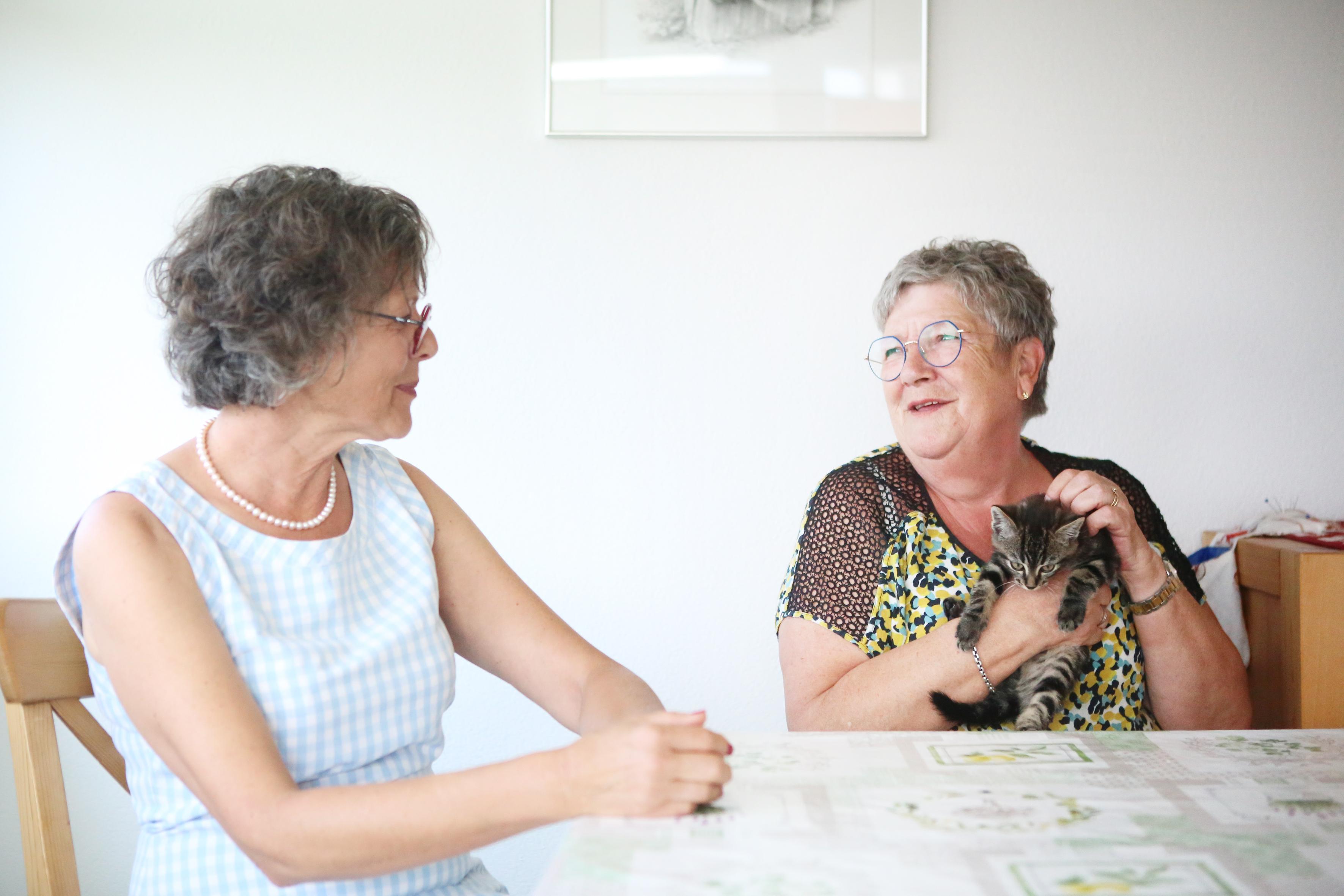 Martine Pipoz und Imelda Morard sitzen  an einem Tisch und unterhalten sich. Imelda Morard streichelt währenddessen eine kleine Katze, die sie in den Armen hält. 