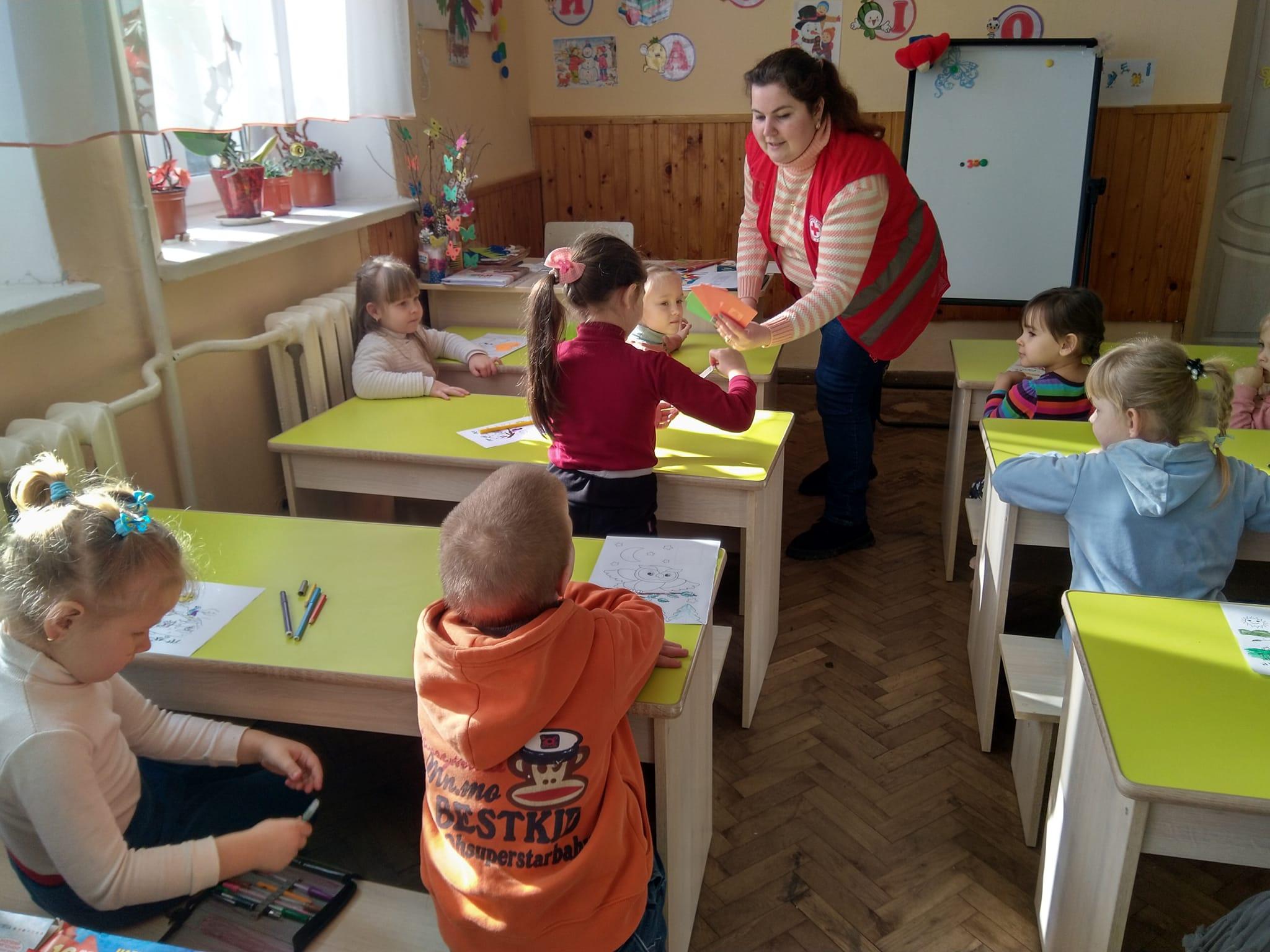 Mehrere Kinder sitzen in einem Klassenzimmer. Eine Frau verteilt Karten zum spielen.