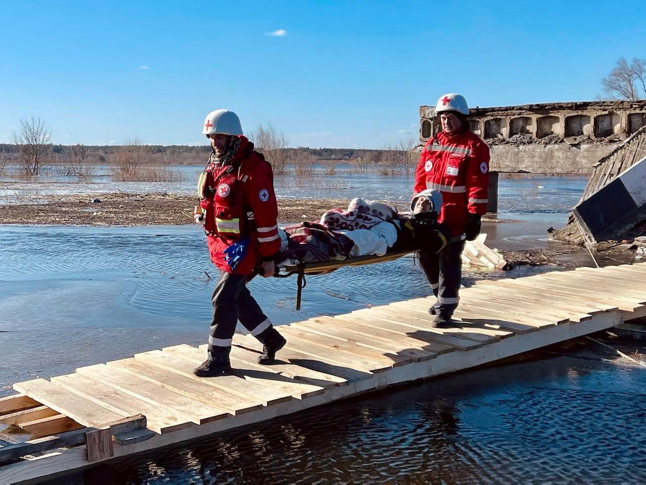 Zwei Freiwillige des ukrainischen Roten Kreuzes evakuieren eine verletzte Person, die mit einer Decke auf einer Trage liegt. Sie überqueren eine provisorische Holzbrücke. Die beiden Freiwilligen tragen den weißen Helm und die rote Jacke des Roten Kreuzes.