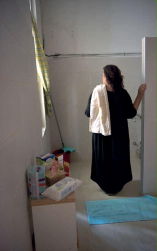 Une femme photographiée de dos, debout dans une salle de bain, une serviette éponge sur l'épaule.