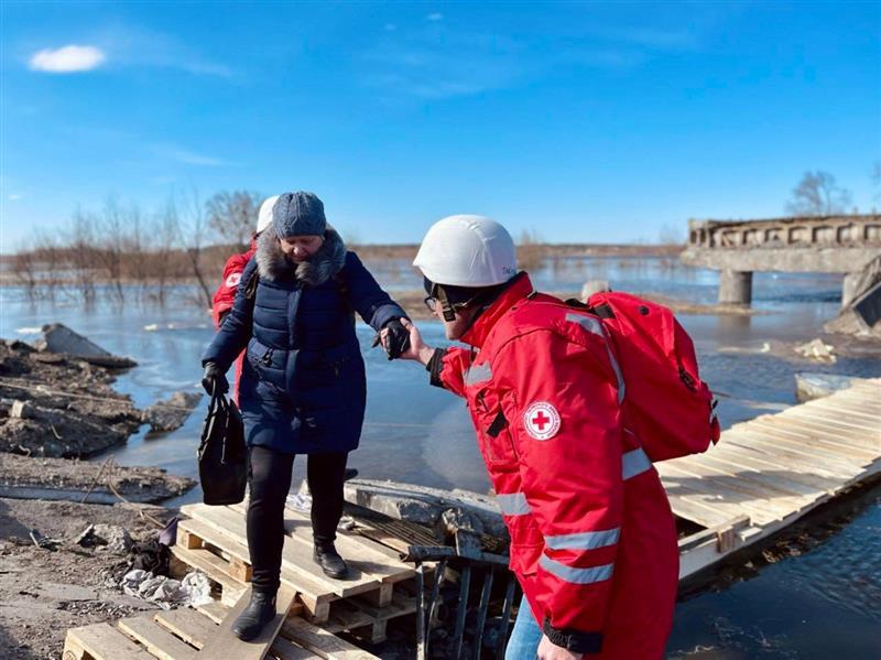 Ein Freiwilliger des Ukrainischen Roten Kreuzes hilft eine Person eine provisorische Holzbrücke zu überqueren. Der Freiwilliger tragt den weißen Helm und die rote Jacke des Ukrainischen Roten Kreuzes.