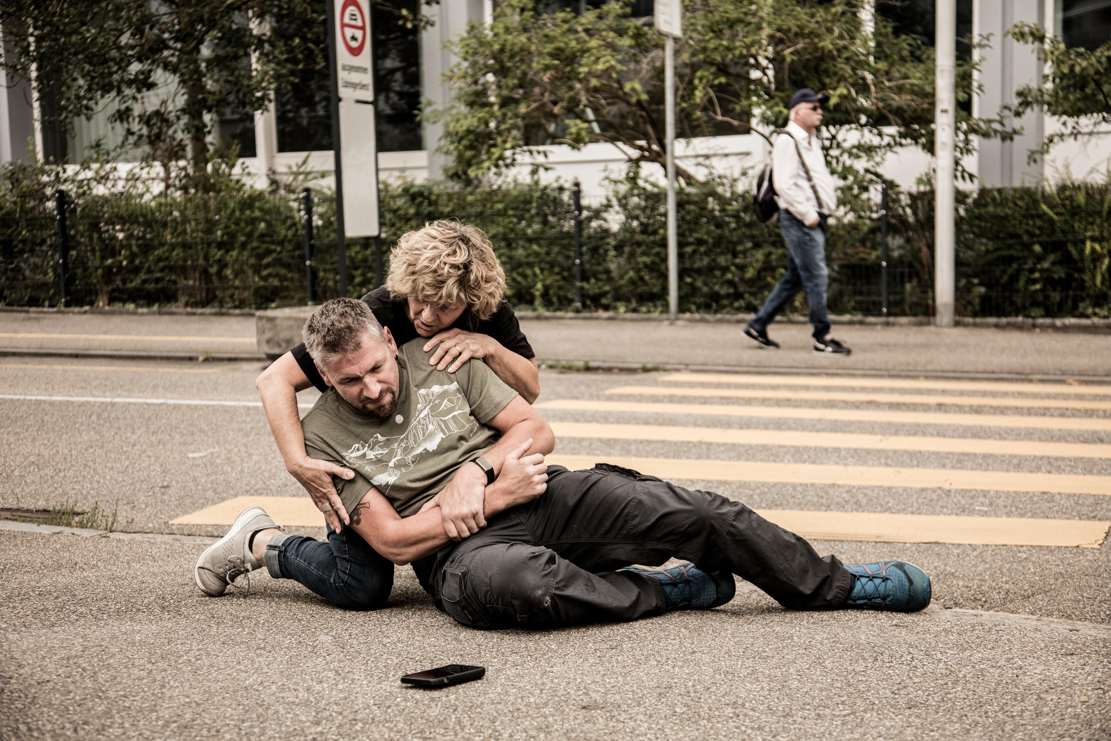 Un homme est tombé. Blessé, il est allongé sur le sol. Une passante s’occupe de lui et lui prodigue les premiers soins.