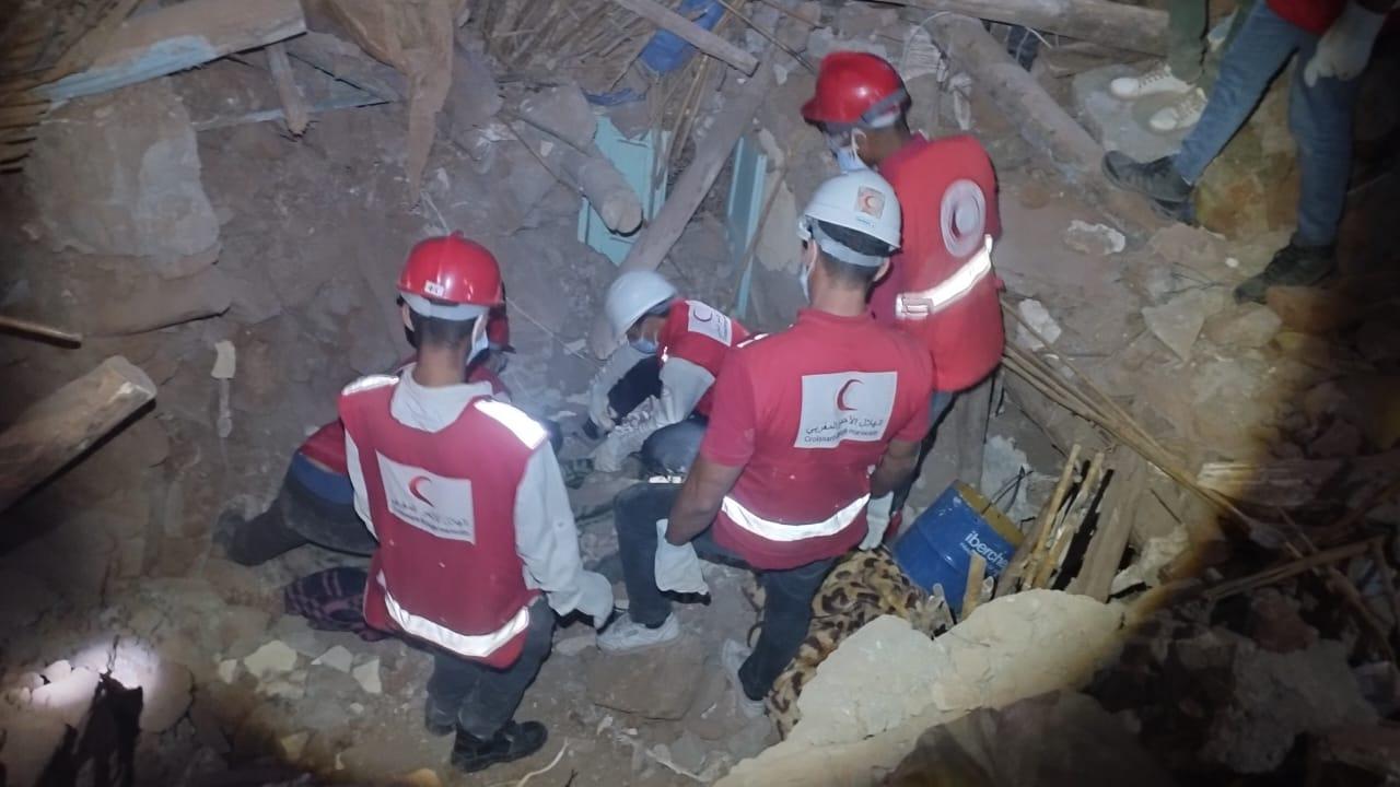 Un groupe de cinq hommes avec des gilets rouge et le logo du Croissant Rouge saont dans les débris d'une maison.