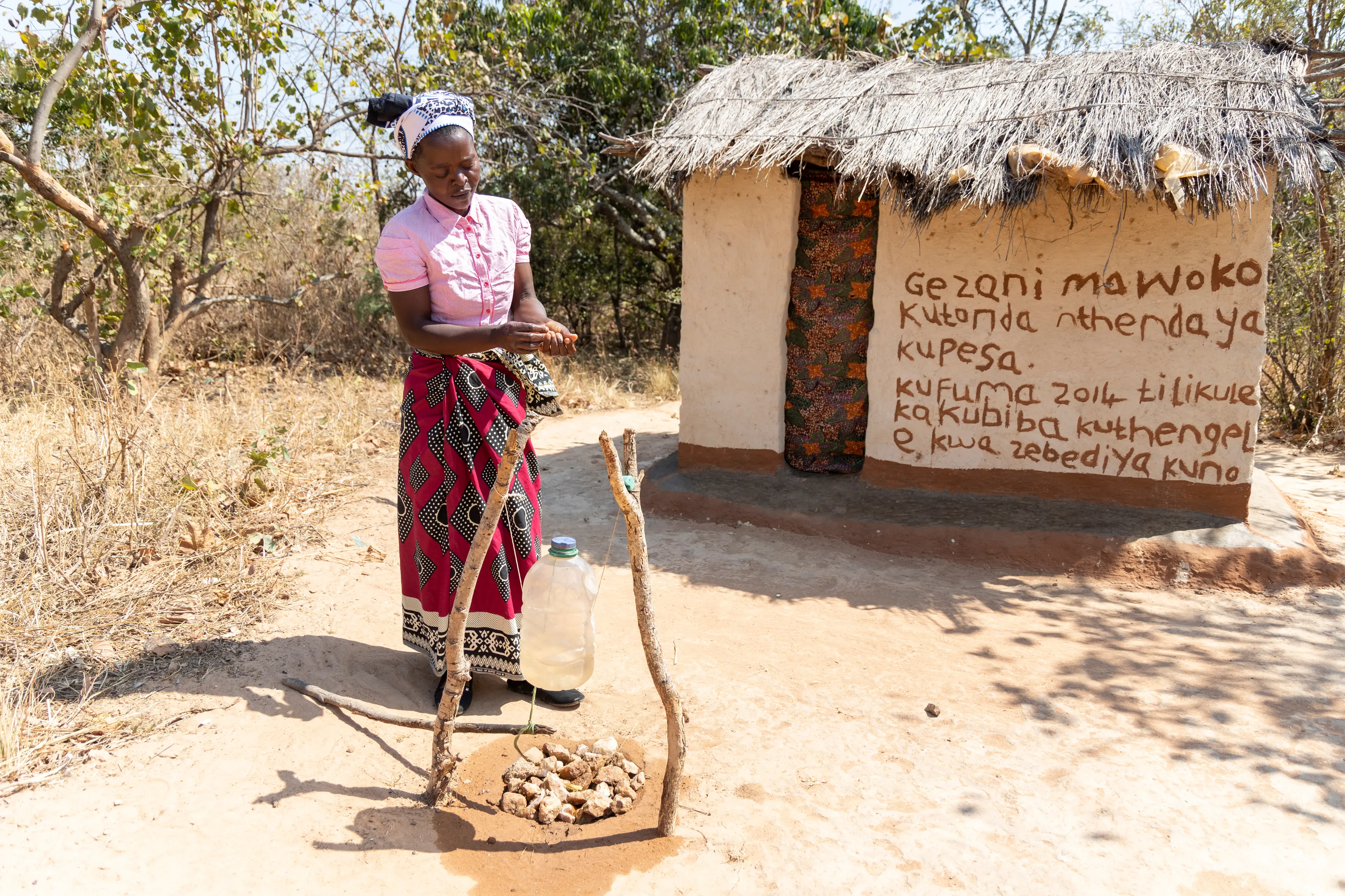 Une femme se lave les mains au Tippy Tap, derrière une hutte en terre et un environnement sec.