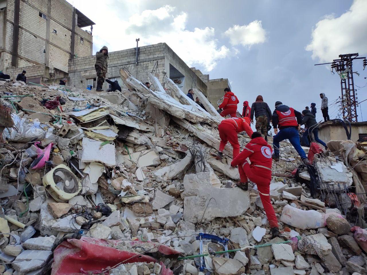 Several men climb a gigantic pile of rubble. Some wear the red pants and jackets of the Turkish Red Crescent. 