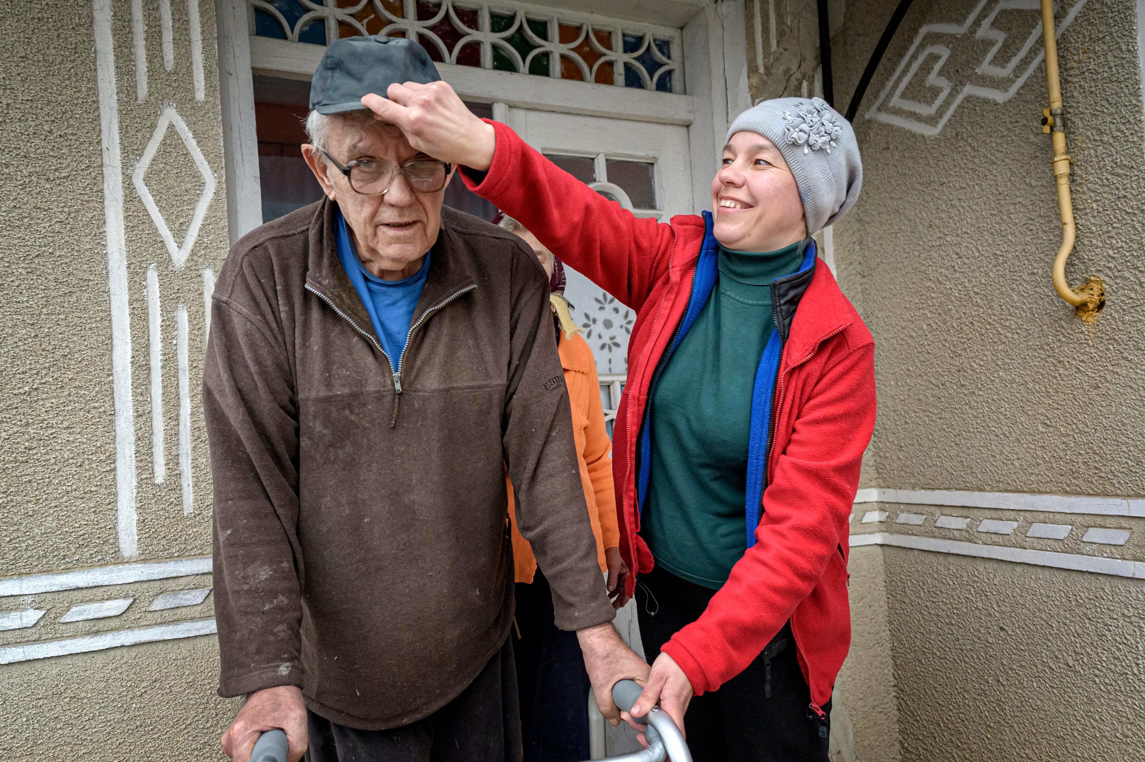 Un uomo anziano è in piedi all'ingresso della casa. Una donna con una giacca rossa gli mette il cappello in testa e ride.