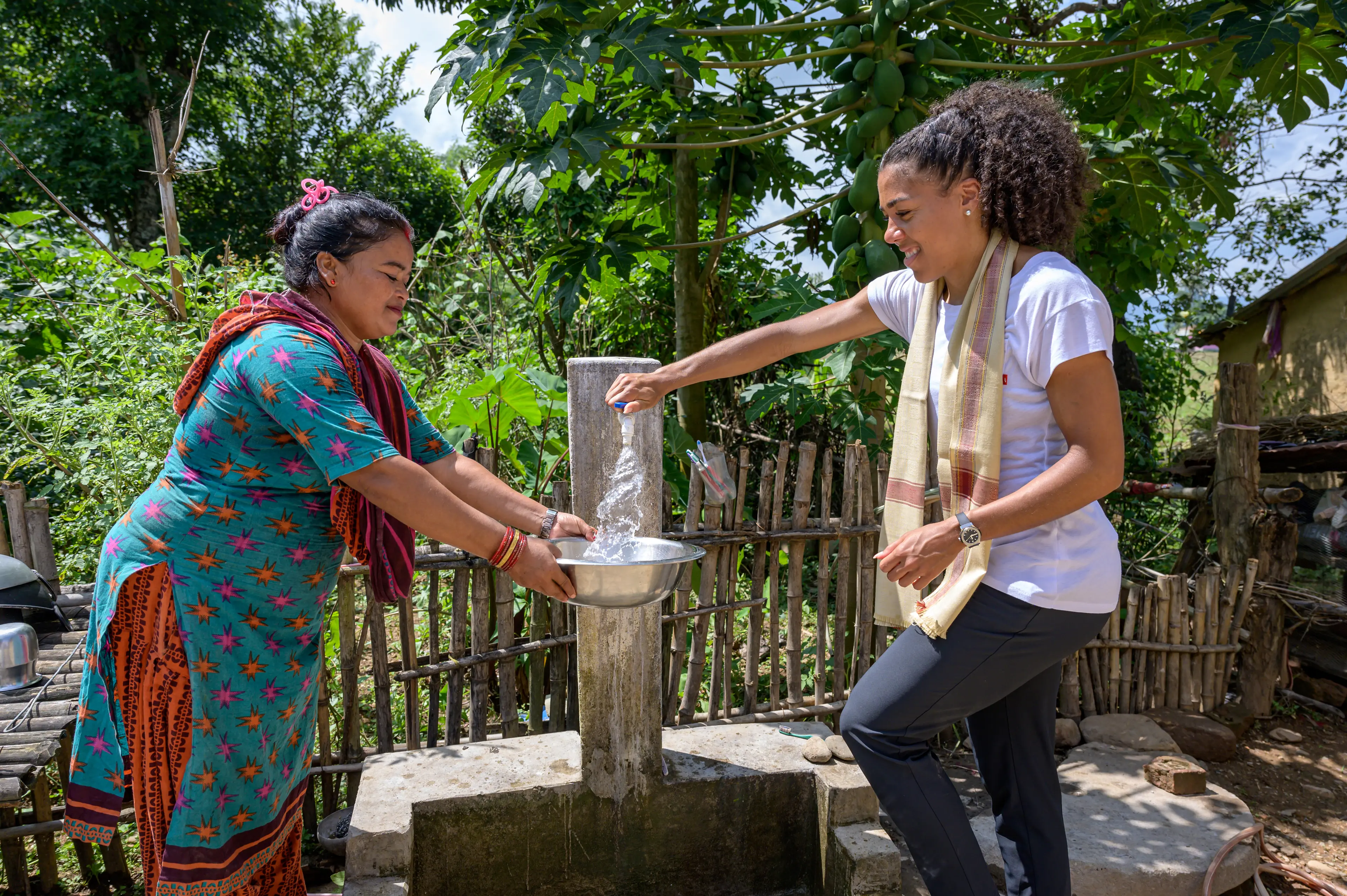 Eine bunt gekleidete Frau und Mujinga Kambundji stehen um einen Wasserhahn im Freien. Mujinga Kambundji hat eine Hand auf dem Wasserhahn, aus dem Wasser fließt. Im Hintergrund ein grüner Garten