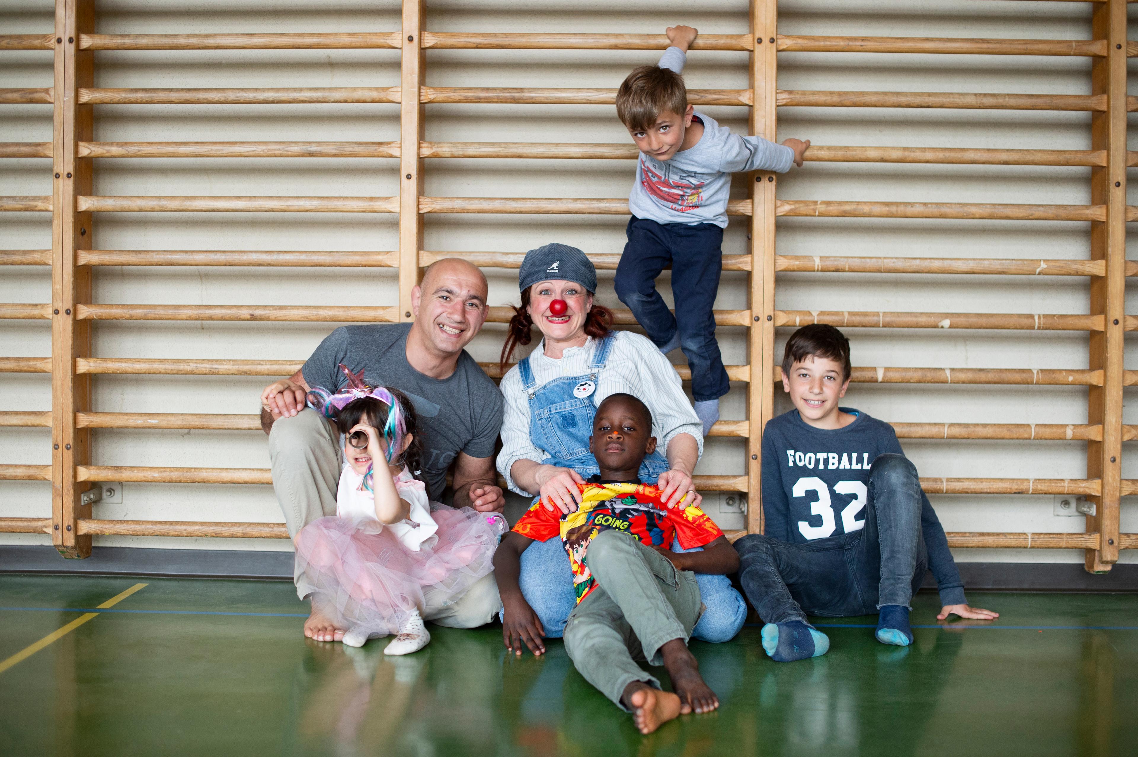 Die vierjährige Marianne, ihr Vater, Clownin Zilibii und zwei Knaben sitzen in einer Turnhalle auf dem Boden.  Ein Knabe hält sich darüber an der Sprossenfwand fest. Alle lachen.