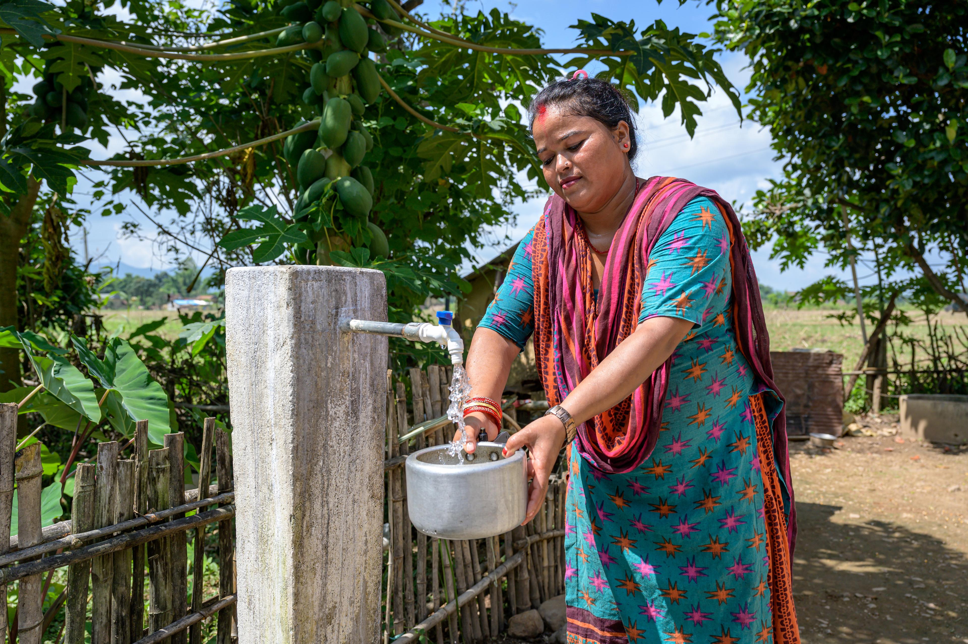 Eine bunt gekleidete Frau bedient in einem Garten im Freien einen Wasserhahn. Sie füllt ein Gefäss, mit Wasser. Im Hintergrund ein grüner Garten.