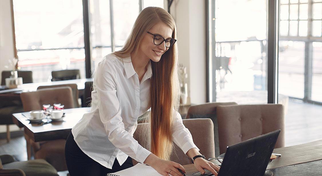 Das Bild zeigt eine Person mit langen, glatten Haaren, die sich &uuml;ber einen Tisch in einem hellen Caf&eacute; oder Arbeitsbereich beugt. Sie tr&auml;gt ein wei&szlig;es Hemd mit Kn&ouml;pfen und arbeitet anscheinend an einem Laptop, mit einem Notizbuch und Stift in der N&auml;he. Im Hintergrund sind gro&szlig;e Fenster zu sehen, die nat&uuml;rliches Licht hereinlassen, mit Tischen und St&uuml;hlen in einem gem&uuml;tlichen, modernen Stil angeordnet. Auf einigen Tischen stehen Tassen und Untertassen, die zur entspannten und dennoch professionellen Atmosph&auml;re beitragen.