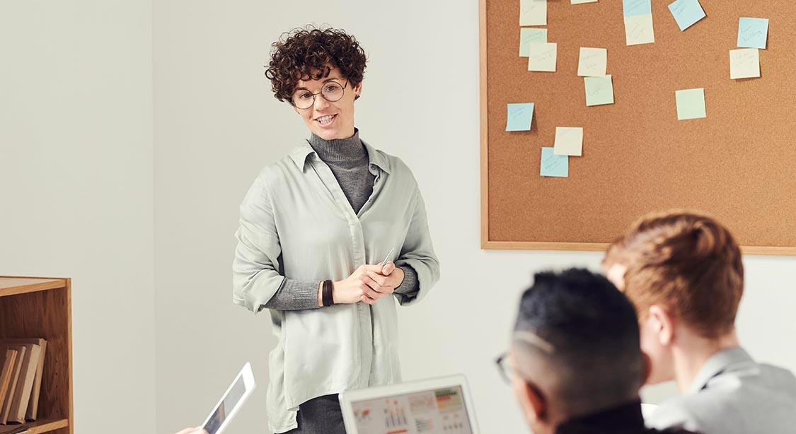 A person is standing at the front of a meeting room and talking with a colleague that is seated with other people at a table.