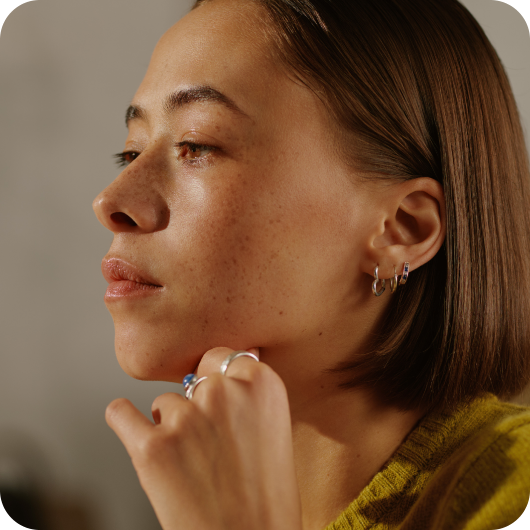 Eine Nahaufnahme einer Person mit glattem, mittellangem braunem Haar, die mehrere kleine Kreolen an einem Ohr tr&auml;gt. Die Person h&auml;lt ihre Hand nah am Gesicht und zeigt dabei einen silbernen Ring. Sie tr&auml;gt einen senfgelben Strickpullover vor einem dezent verschwommenen Hintergrund.