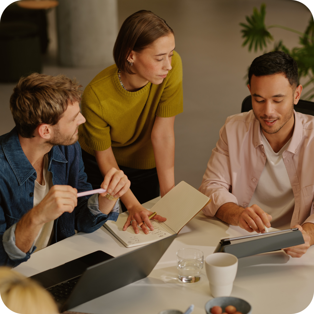 Eine Gruppe von drei Personen, die in einem B&uuml;ro zusammenarbeiten. Eine Person in einem senfgelben Pullover lehnt sich &uuml;ber einen Tisch, h&auml;lt ein Notizbuch und einen Bleistift und scheint das Gespr&auml;ch zu leiten. Eine andere Person in einem Jeanshemd h&auml;lt einen Stift, w&auml;hrend die dritte Person in einem hellrosa Shirt ein Tablet benutzt. Auf dem Tisch liegen verschiedene Gegenst&auml;nde verstreut, darunter ein Laptop, ein Wasserglas, eine Kaffeetasse und eine kleine Schale. Im Hintergrund sind eine Pflanze und moderne B&uuml;rom&ouml;bel zu sehen.