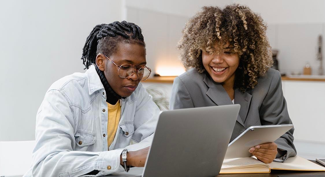 Das Bild zeigt zwei Personen, die in einem modernen, gut beleuchteten Arbeitsbereich zusammenarbeiten. Eine Person tr&auml;gt ein helles Jeanshemd und ein gelbes Oberteil und konzentriert sich auf einen Laptop vor ihr. Die andere Person in einem grauen Blazer h&auml;lt ein Tablet und scheint in das Gespr&auml;ch vertieft zu sein. Auf dem Tisch liegt ein Notizbuch, und im Hintergrund ist ein minimalistisches Interieur mit neutralen Farbt&ouml;nen und dezenter Dekoration zu sehen.