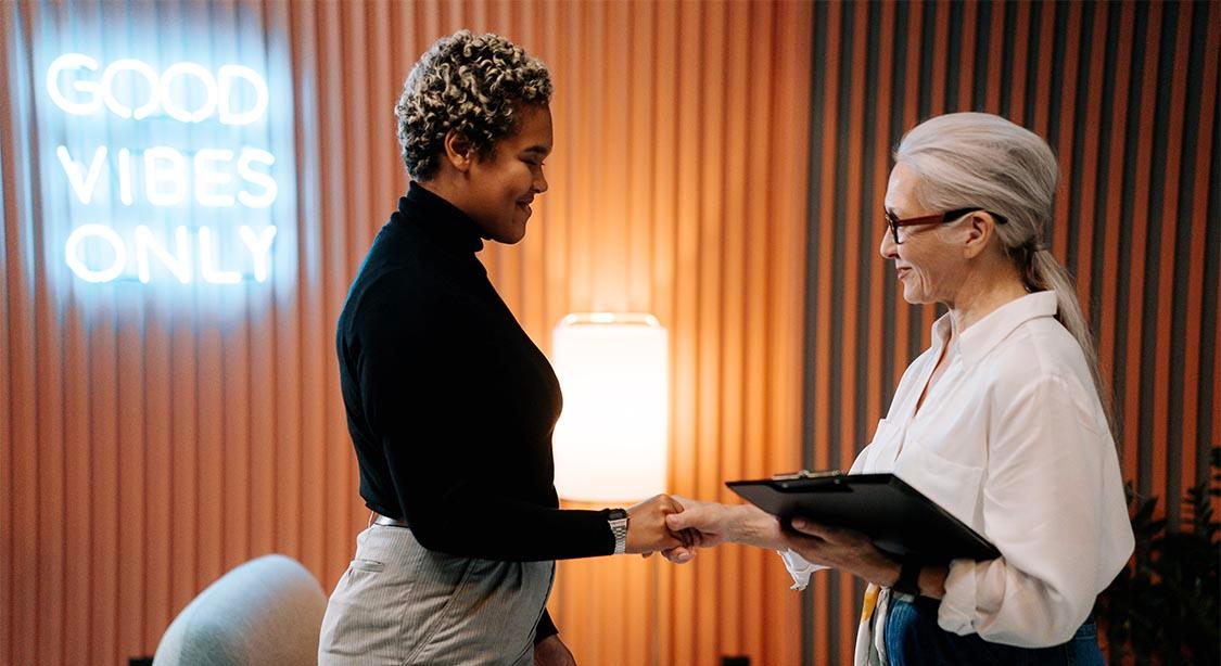 Das Bild zeigt zwei Personen, die sich in einer professionellen Umgebung mit warmer und moderner Atmosph&auml;re die Hand geben. Eine Person tr&auml;gt einen schwarzen Rollkragenpullover und eine graue Hose, w&auml;hrend die andere ein wei&szlig;es Hemd tr&auml;gt und ein Klemmbrett h&auml;lt. Im Hintergrund sind vertikale Holzpaneele und ein leuchtendes Neonschild mit der Aufschrift "GOOD VIBES ONLY" zu sehen, zusammen mit einer eingeschalteten Stehlampe, die eine einladende Atmosph&auml;re schaffen.