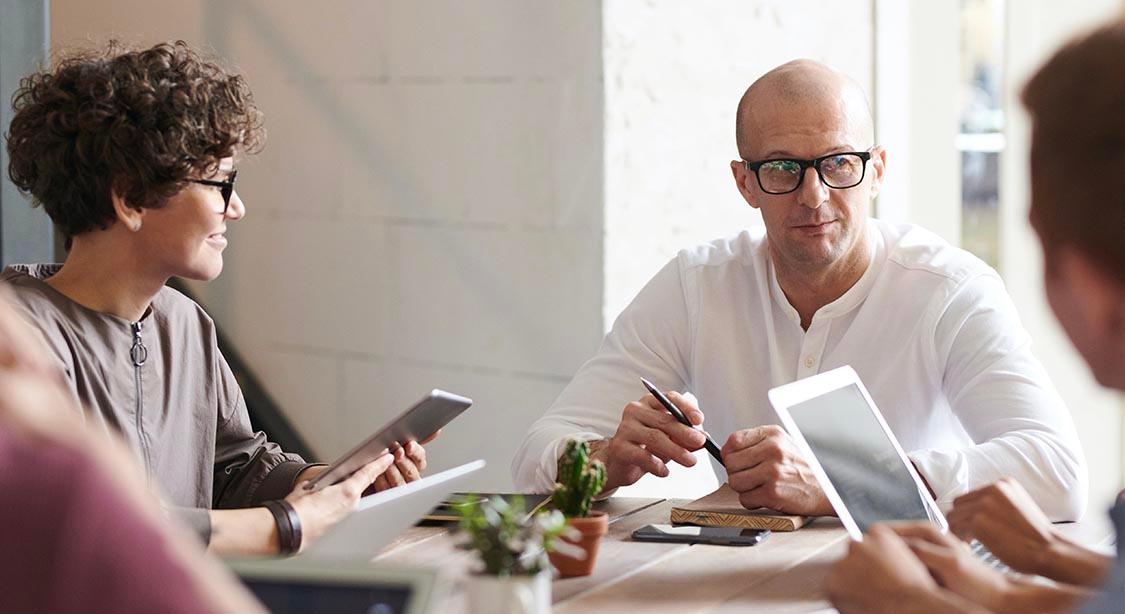 Eine Gruppe von Personen sitzt um einen Holztisch in einer hellen, modernen B&uuml;roumgebung. Sie sind in ein Gespr&auml;ch vertieft, wobei einige Tablets und Notizb&uuml;cher in den H&auml;nden halten. Auf dem Tisch steht eine kleine Topfpflanze, die dem Arbeitsbereich einen Hauch von Gr&uuml;n verleiht.